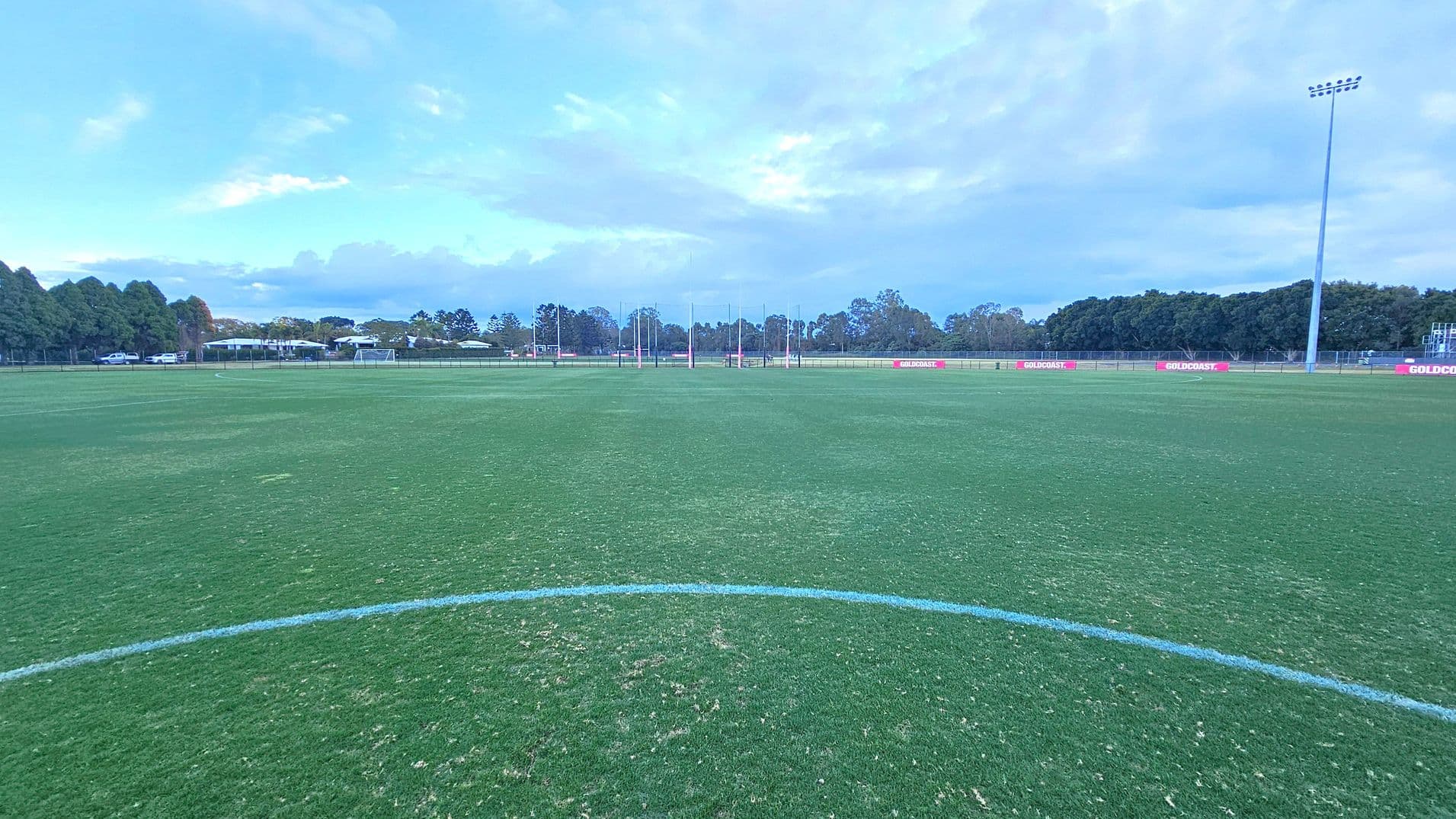 A large, open sports field with well-maintained grass. In the centre, there are goalposts in the distance. The foreground shows a curved white line on the grass. To the right, a tall floodlight pole stands near the edge of the field. The background features a line of trees and some buildings, with a few parked cars visible. Red signs with "GOLDCOAST" are placed along the perimeter fence.