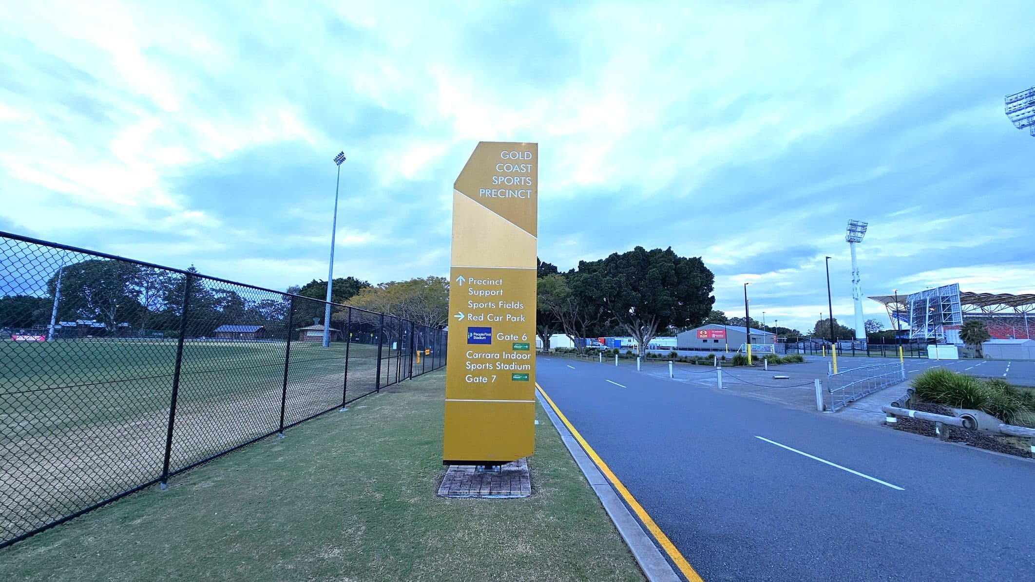 A tall, yellow sign stands on a grassy area, displaying directions for the Gold Coast Sports Precinct. To the left, a chain-link fence encloses a sports field with trees in the background. On the right, a paved road runs alongside the sign, leading towards a stadium with visible floodlights and seating. The area is bordered by trees and some low buildings.