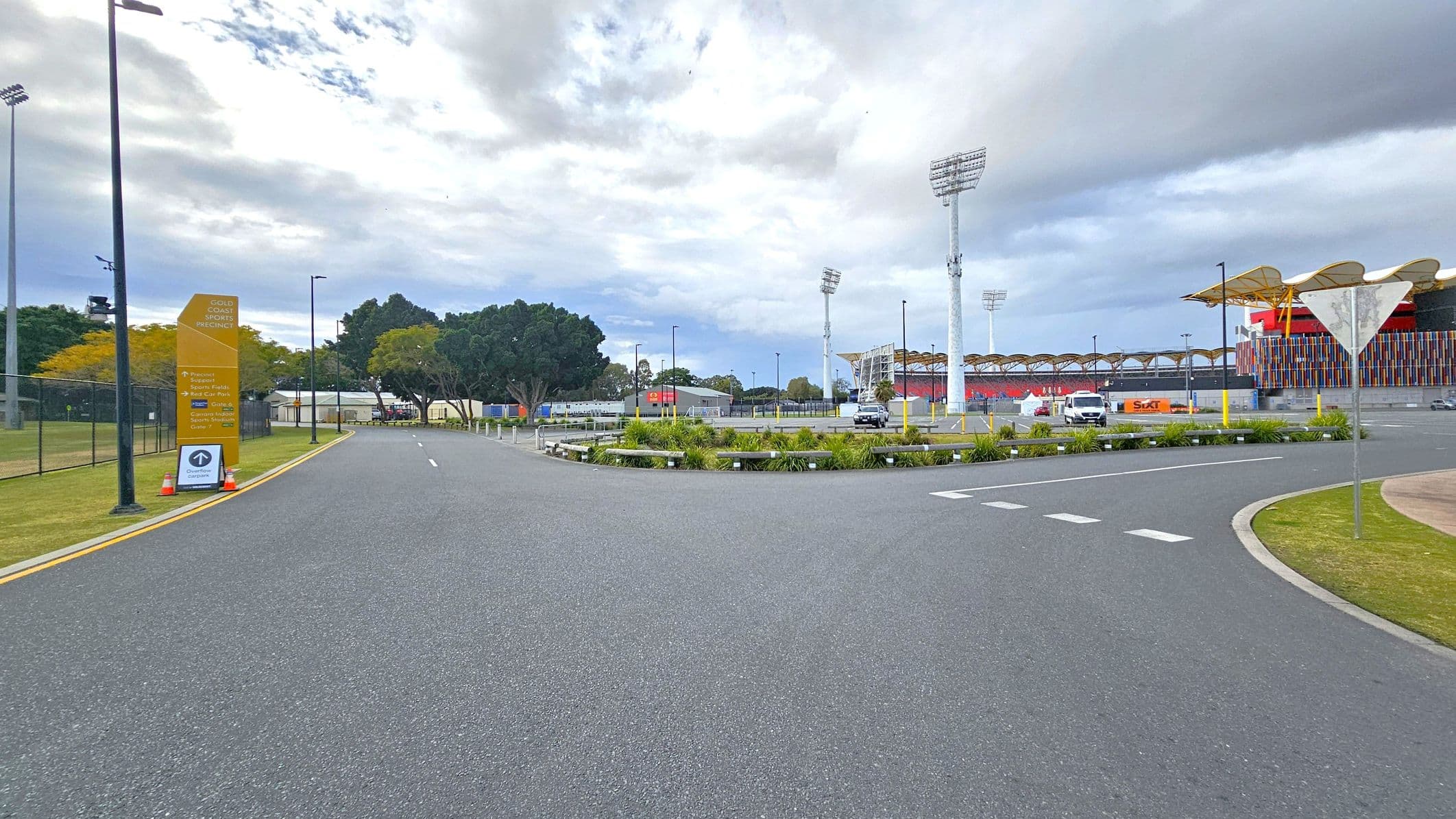 A wide road curves to the right, bordered by a grassy area with a few small plants. On the left, a tall yellow sign reads "Gold Coast Sports Precinct" with additional information below. Several tall floodlights are visible in the background, along with a large stadium structure featuring colourful vertical panels. A few vehicles are parked near the stadium, and trees are scattered throughout the area. The sky is overcast.