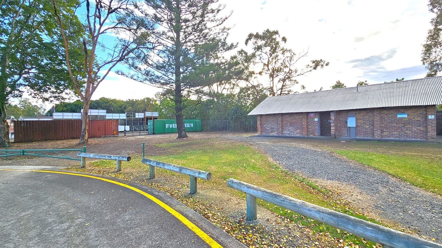 A paved road with a yellow line curves on the left, bordered by wooden barriers. To the right, a long brick building with a corrugated metal roof stands on a gravel path. In the centre, a large tree is surrounded by grass and scattered leaves. Behind the tree, shipping containers, including a green one marked "EVERGREEN," are visible. Tall trees form a backdrop, with a mix of green and brown foliage.