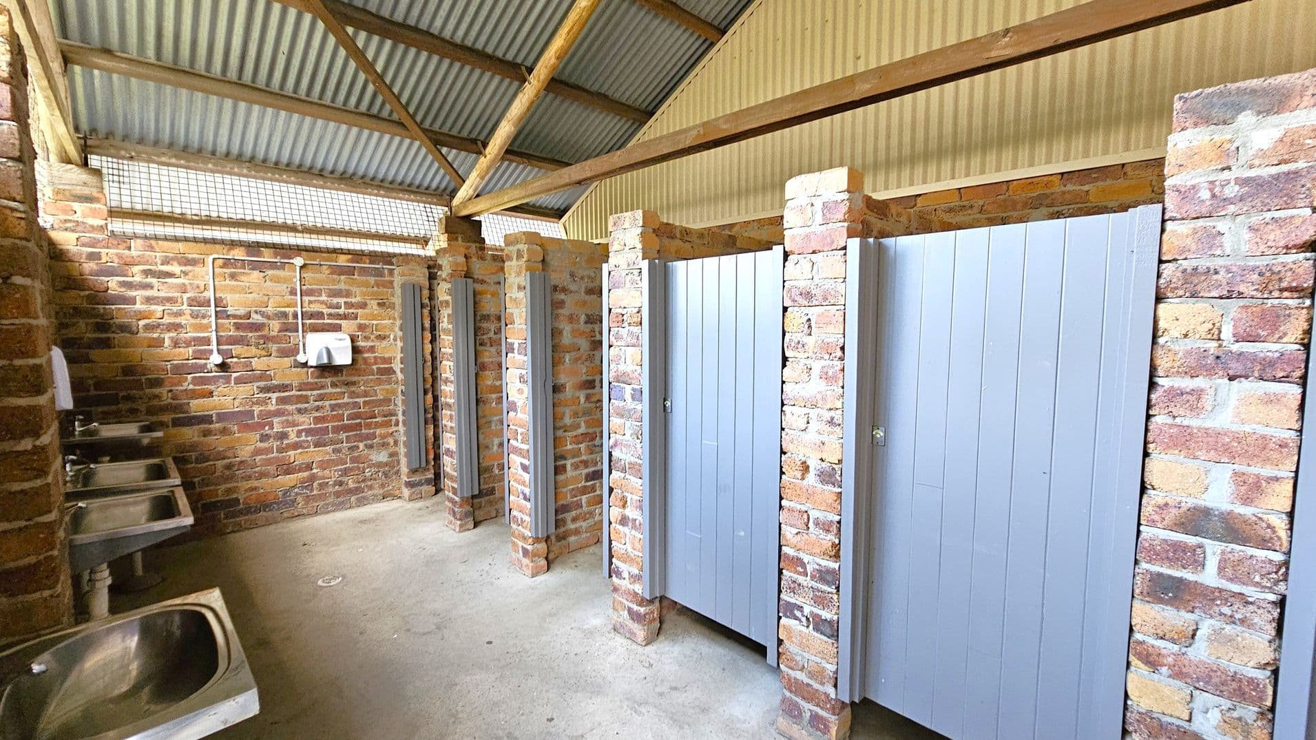 A public restroom with brick walls and a corrugated metal roof. On the left, there are several stainless steel sinks with a paper towel dispenser above. On the right, there are toilet cubicles with grey doors, separated by brick pillars. The floor is concrete, and the ceiling has exposed wooden beams.