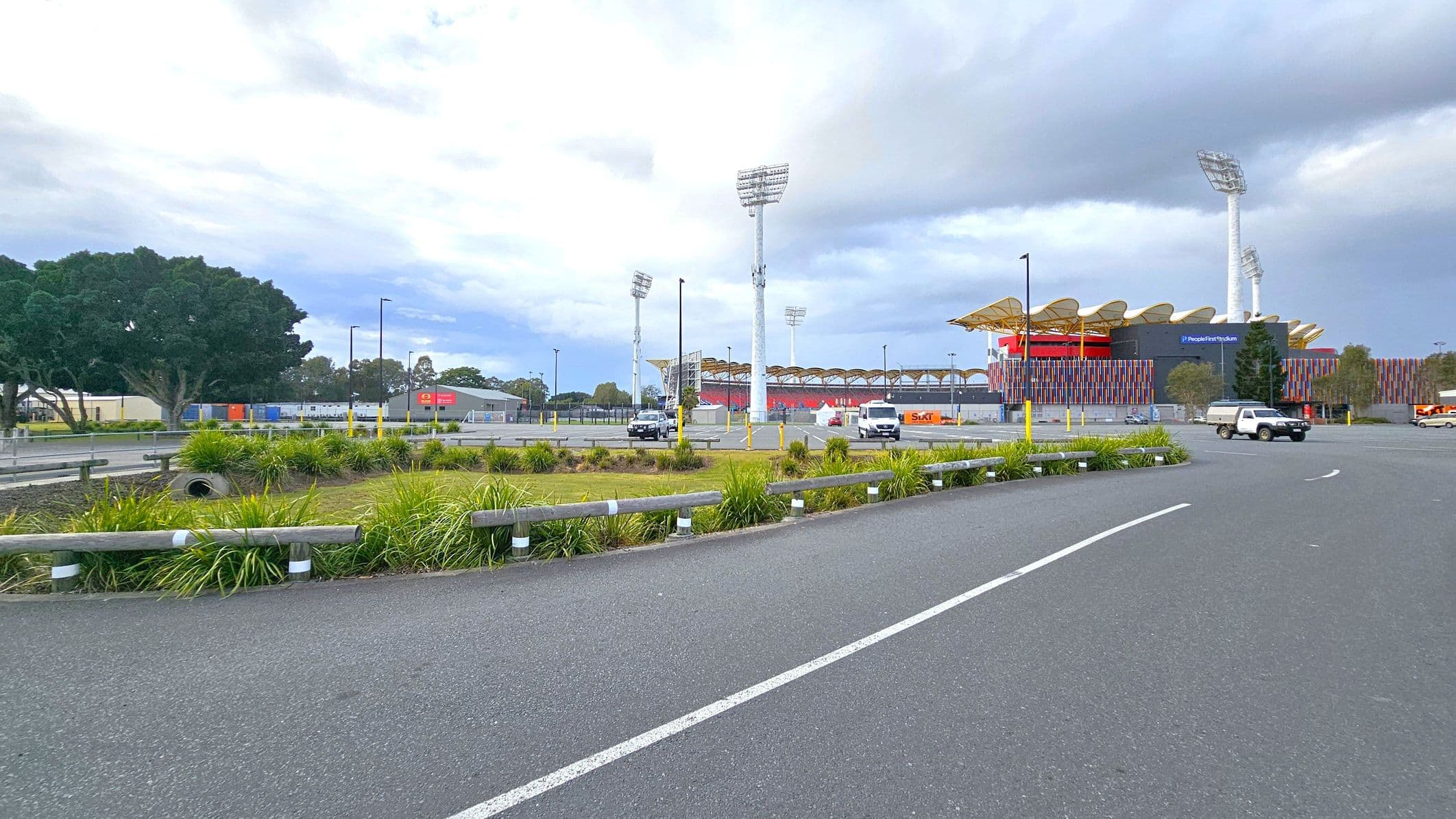 A large stadium with colourful vertical panels and a wavy roof is on the right, with tall floodlights surrounding it. In the foreground, there is a paved road with white lines and a grassy area with low bushes and a metal guardrail. Several vehicles, including a white van and a black car, are parked near the stadium. On the left, there are large trees and some industrial buildings with signage. The sky is overcast.