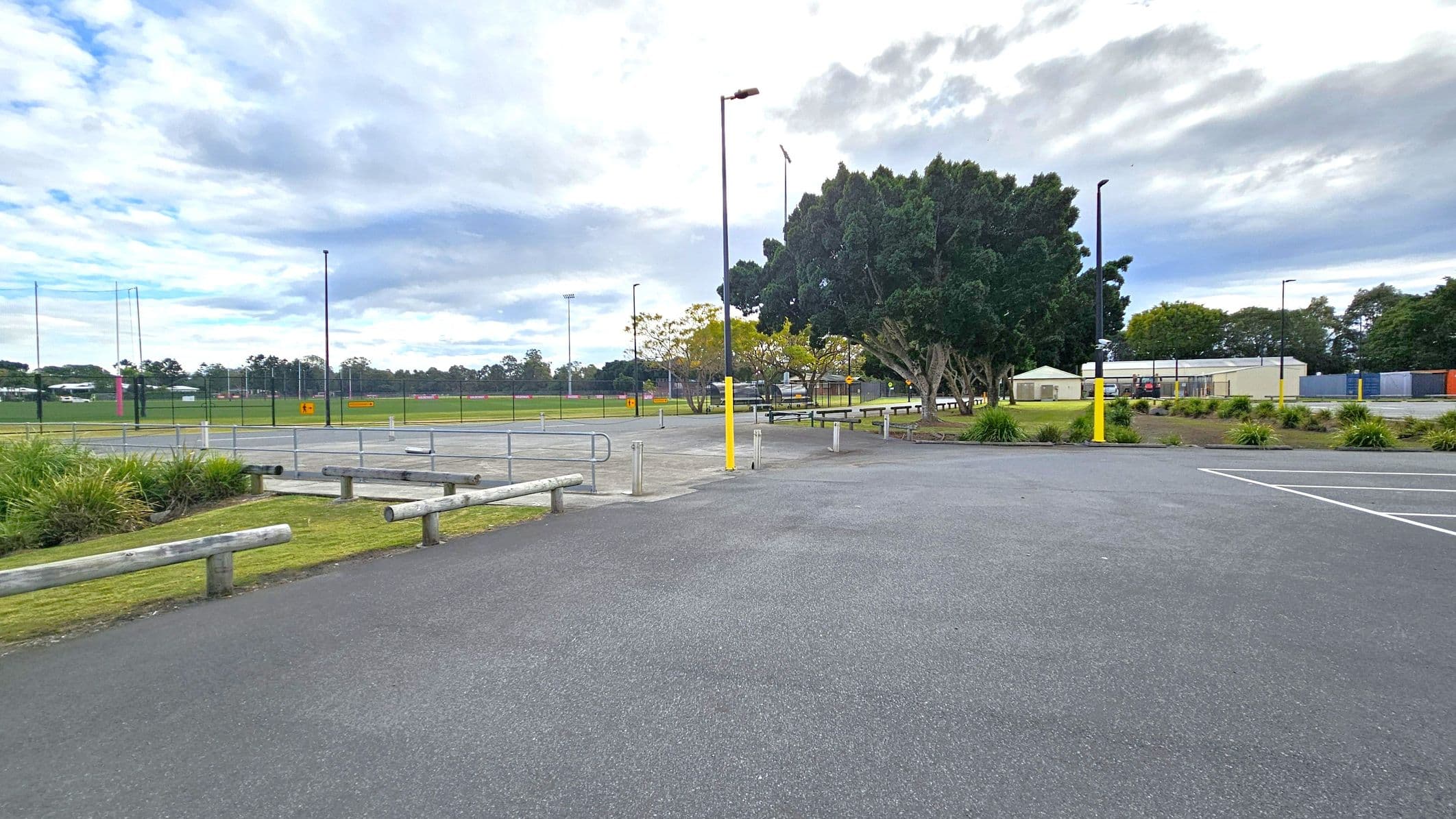 A paved parking area with marked spaces in the foreground. On the left, there are wooden barriers and a grassy area with plants. In the middle, a few tall lamp posts and a fenced sports field with goalposts are visible. On the right, several trees and a few small buildings, possibly storage units or sheds, are present. The background features more trees and a cloudy sky.