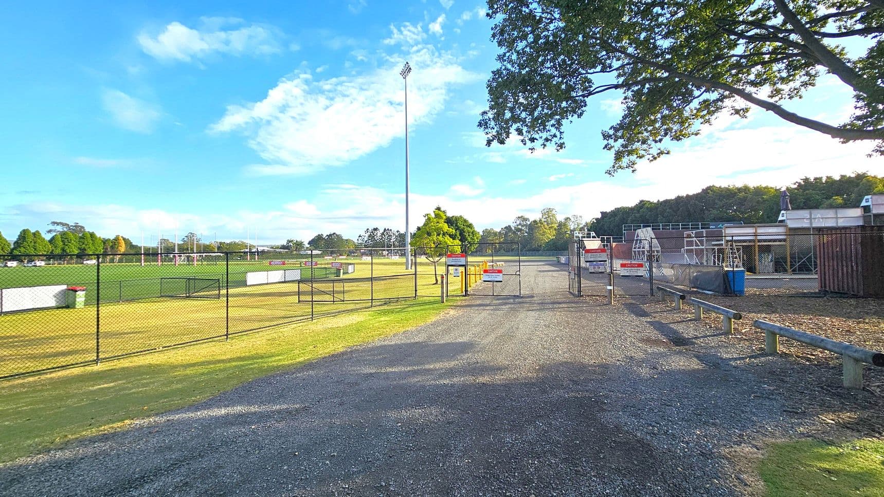 A gravel path leads to a fenced area with a gate in the centre. On the left, there's a green sports field with goalposts and a tall floodlight. A chain-link fence separates the field from the path. On the right, there are structures resembling bleachers or seating areas, with a blue bin nearby. Trees are visible in the background, and a large tree with overhanging branches is on the right. The sky is clear with some clouds.