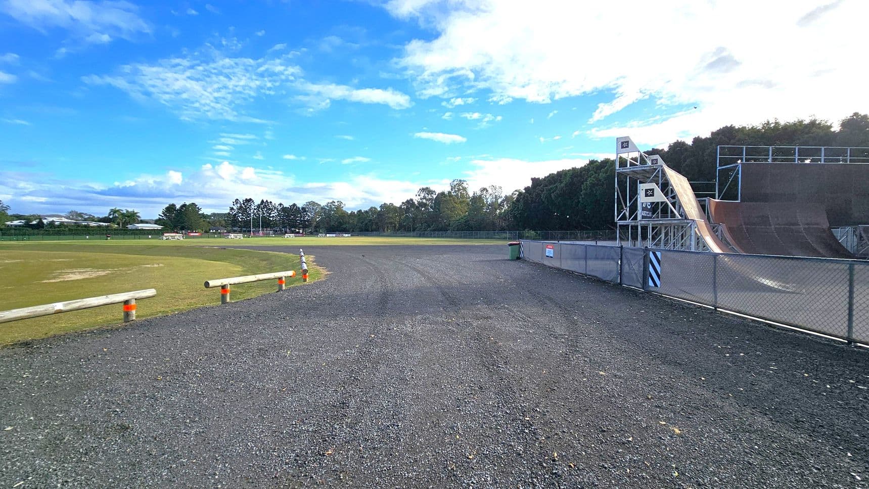 A gravel path runs through the centre, bordered by a grassy field on the left with wooden barriers and orange cones. On the right, a large skate ramp structure with metal railings and platforms is visible, enclosed by a chain-link fence. In the background, there are trees and a few buildings, with a clear blue sky above.