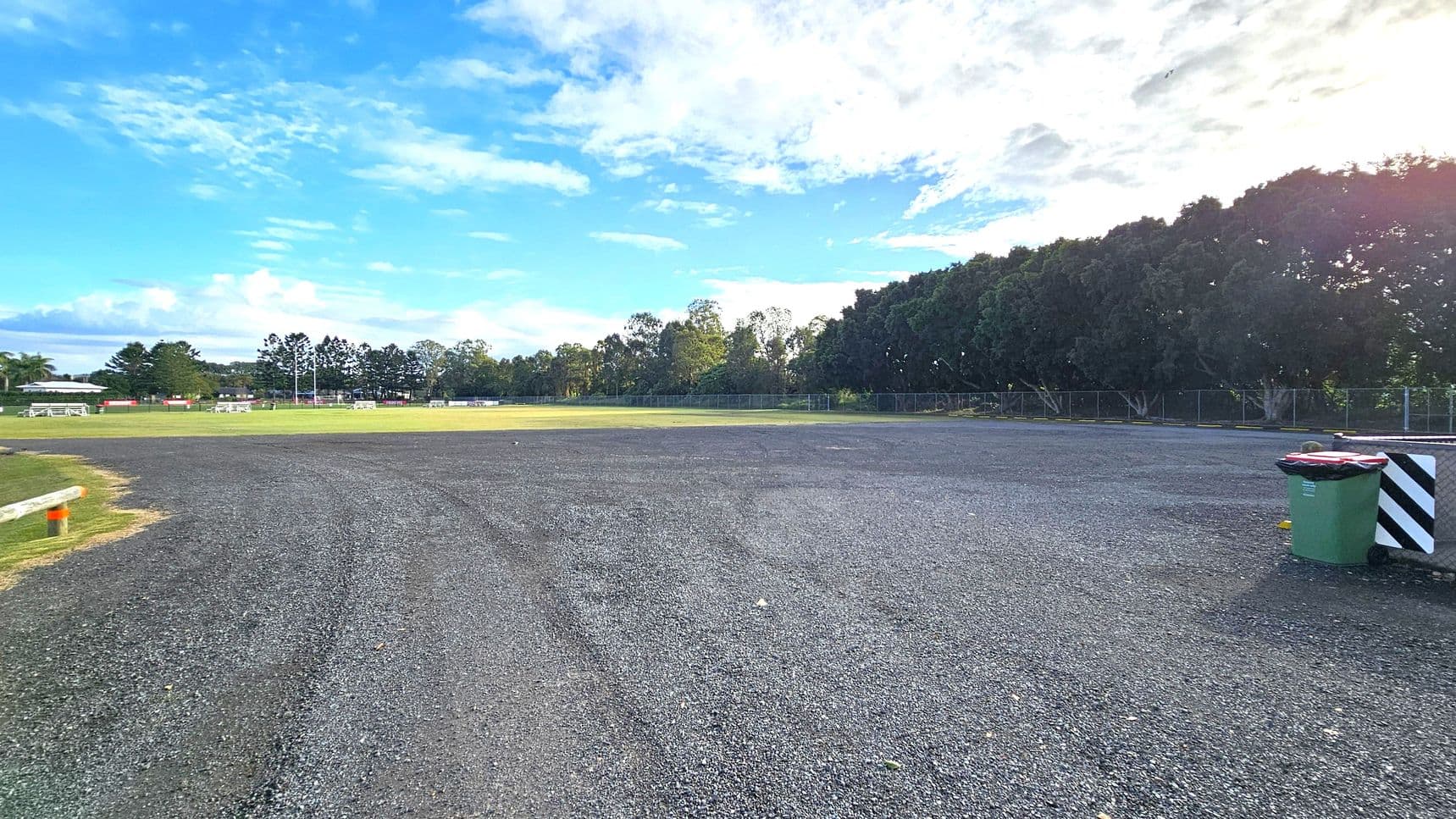 A gravel area in the foreground with a green rubbish bin on the right. In the background, there's a grassy field with white goalposts and benches. Trees line the far edge of the field, and a fence runs along the perimeter. The sky is partly cloudy.