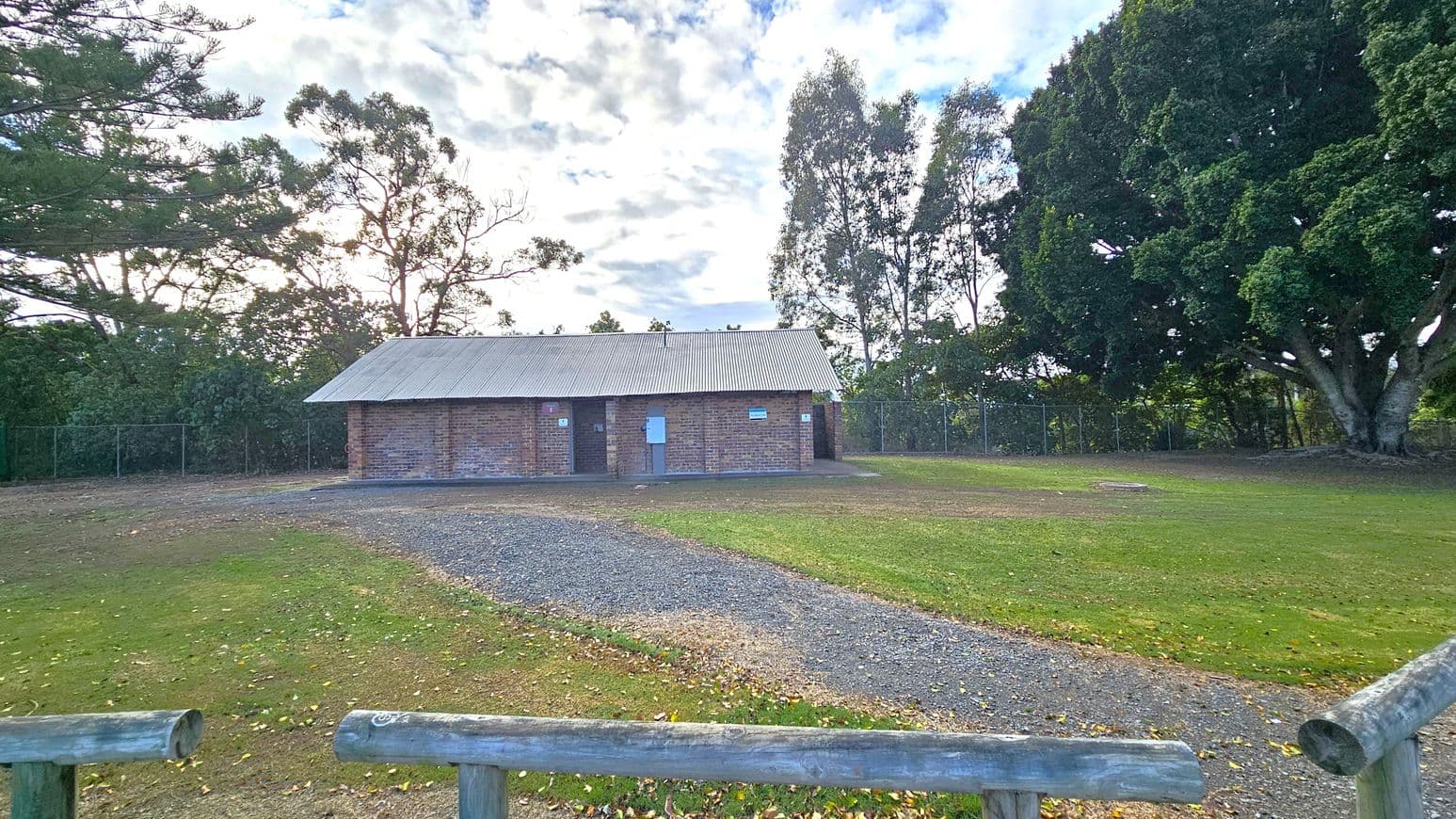 A small brick building with a corrugated metal roof is in the centre, surrounded by a grassy area. A gravel path leads up to the building. On the right, there is a large tree with dense foliage. On the left, there are more trees and a wooden fence in the foreground. The background is filled with more trees and a partly cloudy sky.