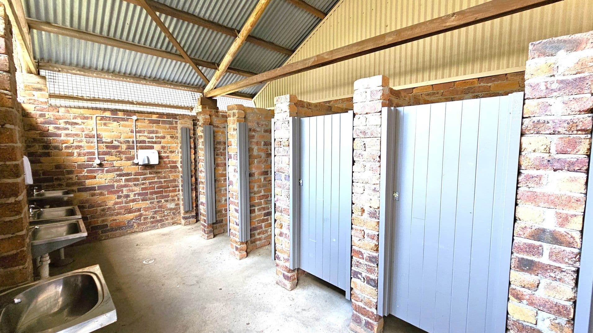 A public restroom with brick walls and a corrugated metal roof. On the left, there are three stainless steel sinks with a soap dispenser and a hand dryer mounted on the wall. On the right, there are several toilet cubicles with light grey doors, separated by brick pillars. The floor is concrete, and the ceiling has exposed wooden beams.