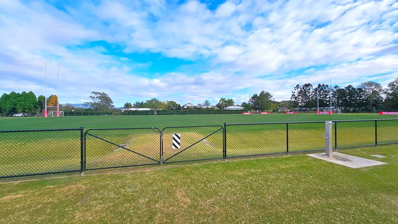 A grassy sports field with goalposts on both the left and right sides. A black chain-link fence runs across the foreground, with a small gate in the centre. Behind the field, there are trees and a few low buildings. The grass is well-maintained, and there are signs with "GOLDCOAST" written on them along the perimeter. The sky is partly cloudy.