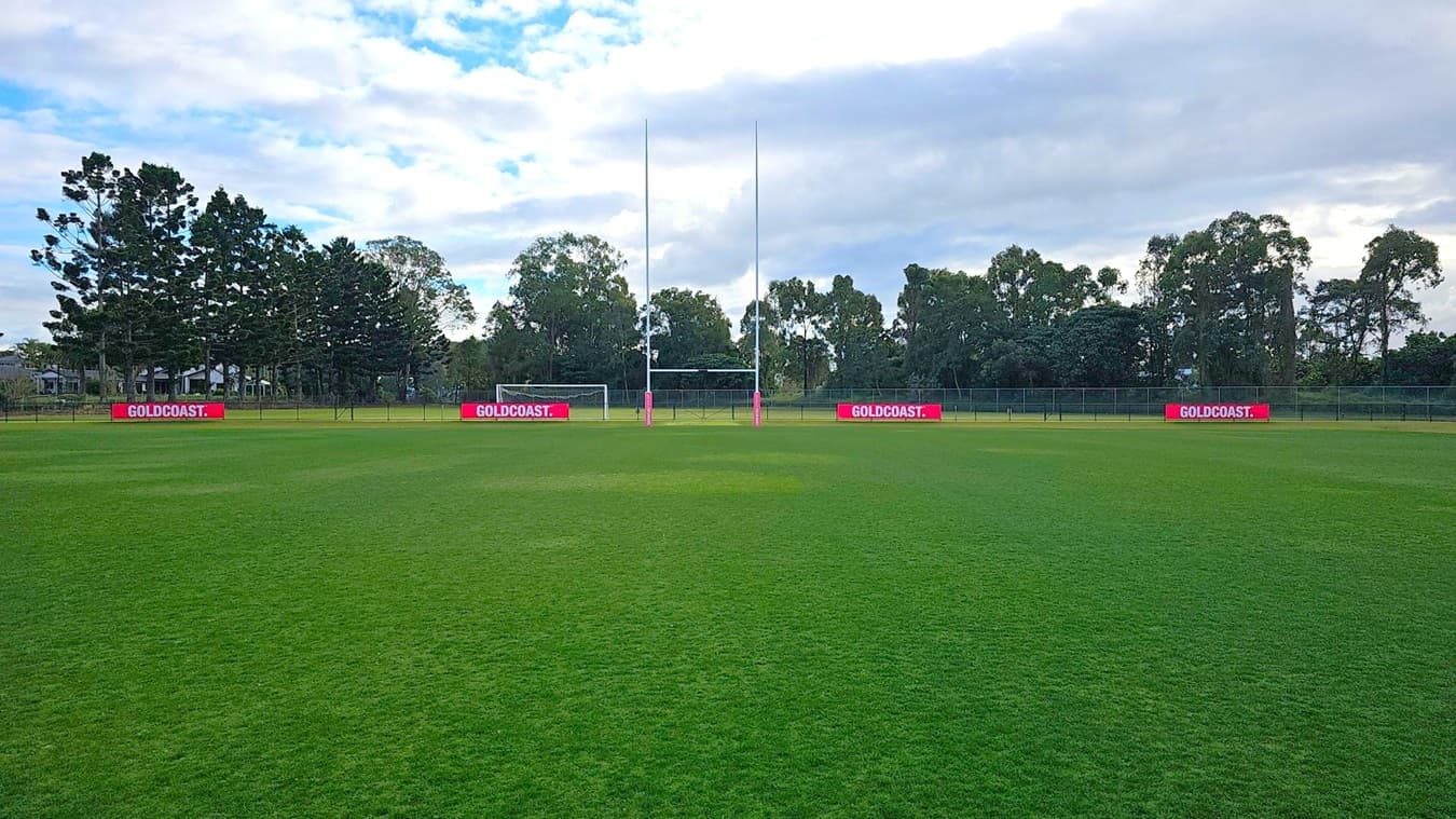 A rugby field with lush green grass. In the centre, there are rugby goalposts with a football goal behind them. Red advertising boards with "GOLDCOAST." are placed along the fence. Tall trees form a dense line in the background, providing a natural border. The sky is partly cloudy, casting a soft light over the scene.