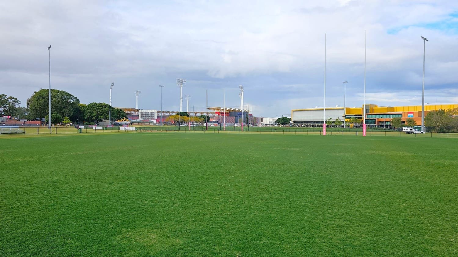 A large, open grassy field with rugby posts in the centre. In the background, there are several tall floodlights and a mix of trees and buildings. On the right, a large yellow and grey building is visible, with a white vehicle parked nearby. The field is surrounded by a low fence, and there are benches and a bin on the left side.