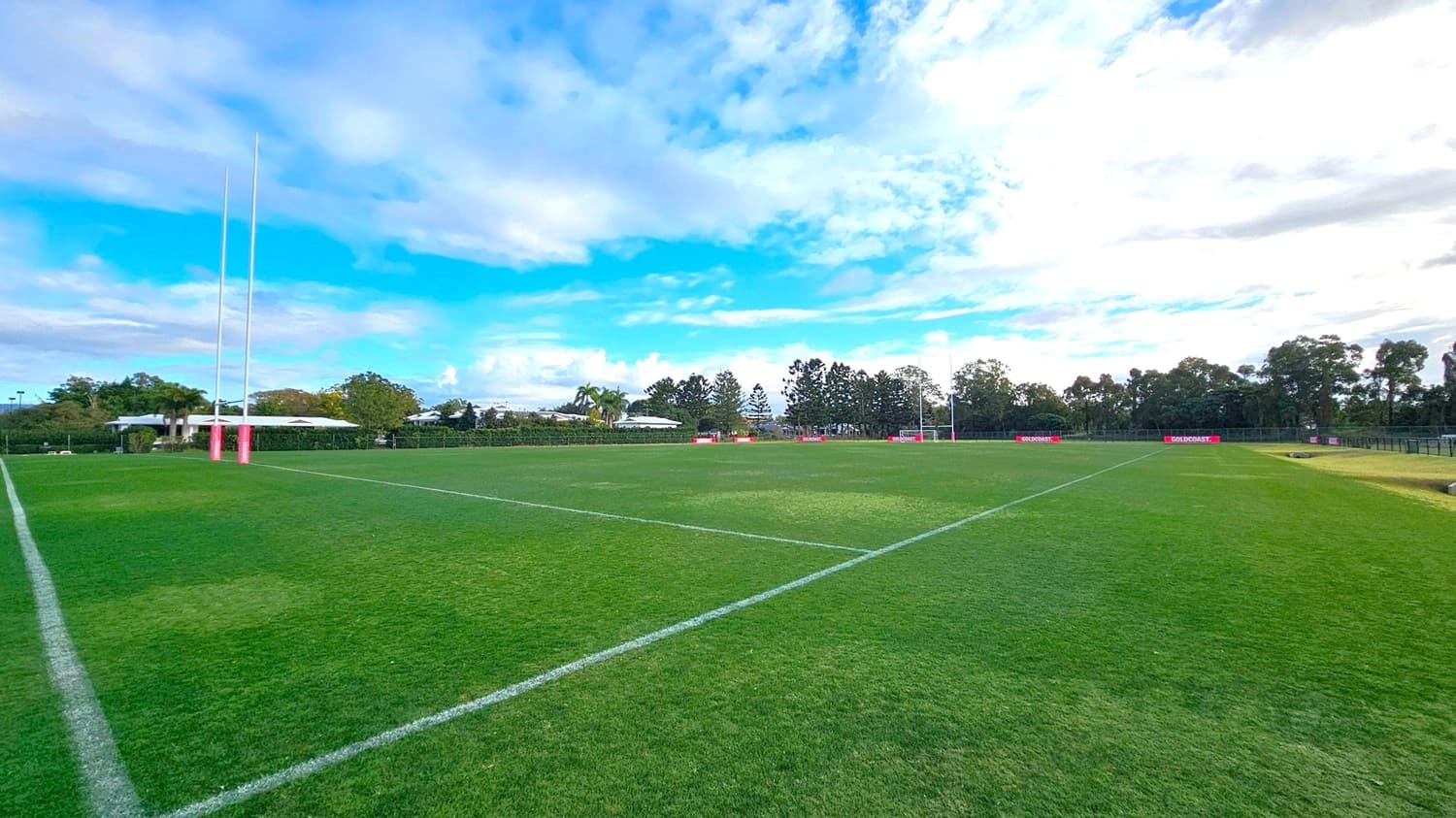 A rugby field with lush green grass and white boundary lines. On the left, there are tall goalposts with pink padding at the base. In the background, there are trees and a few low buildings. On the right, another set of goalposts is visible, and there are red advertising boards along the perimeter. The field is expansive and well-maintained.