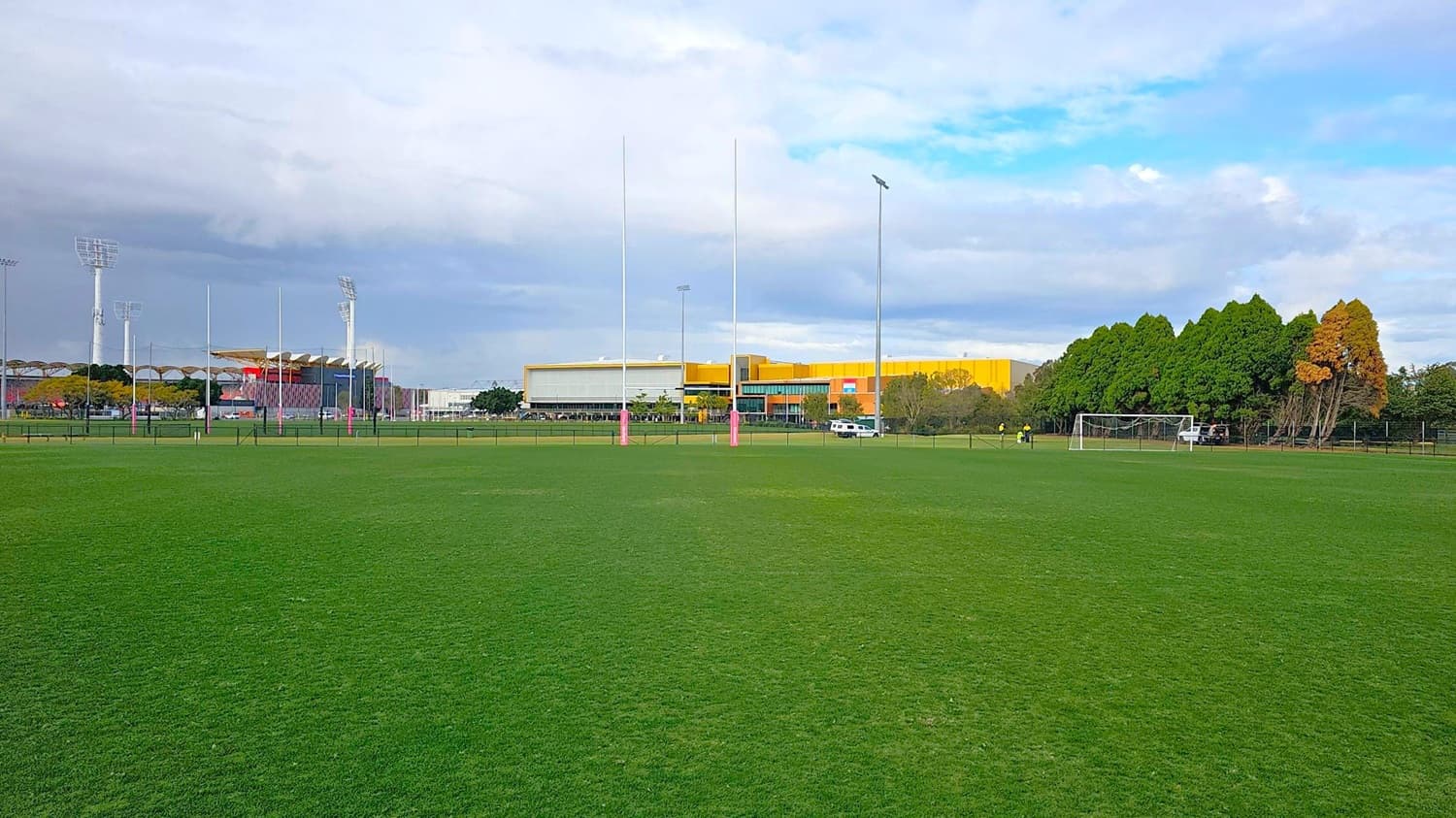 A sports field with green grass in the foreground. In the middle, there are rugby posts with pink padding. To the right, a football goal is visible near a line of trees with some autumn foliage. In the background, there are large buildings with a mix of yellow and grey colours, and several tall floodlights. A white vehicle is parked near the buildings.