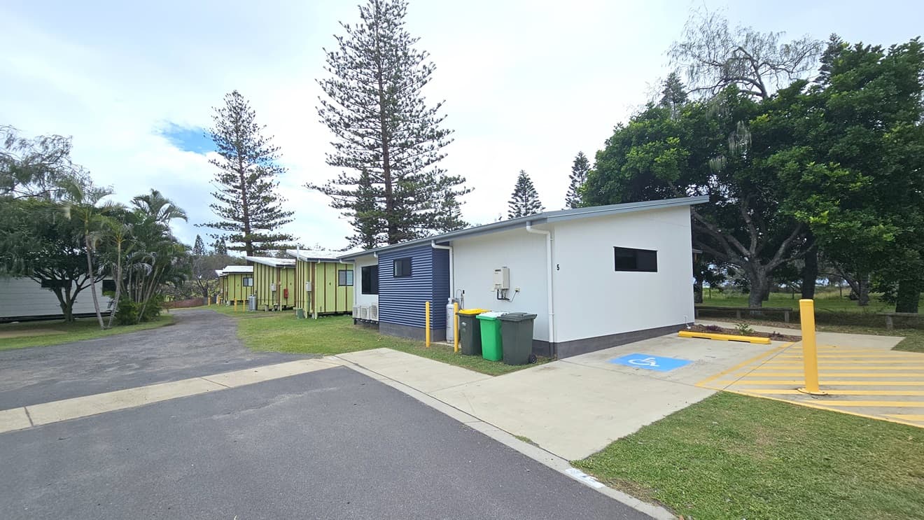 The accessible cabin is nestled on green grass near the park. Accessible parking, marked by a blue wheelchair sign and yellow diagonal lines with a bollard, is on the right. Three rubbish bins are at the back. To the left, the road leads to other cabins, while another facility building is nearby. On the right is a park with pine and palm trees.