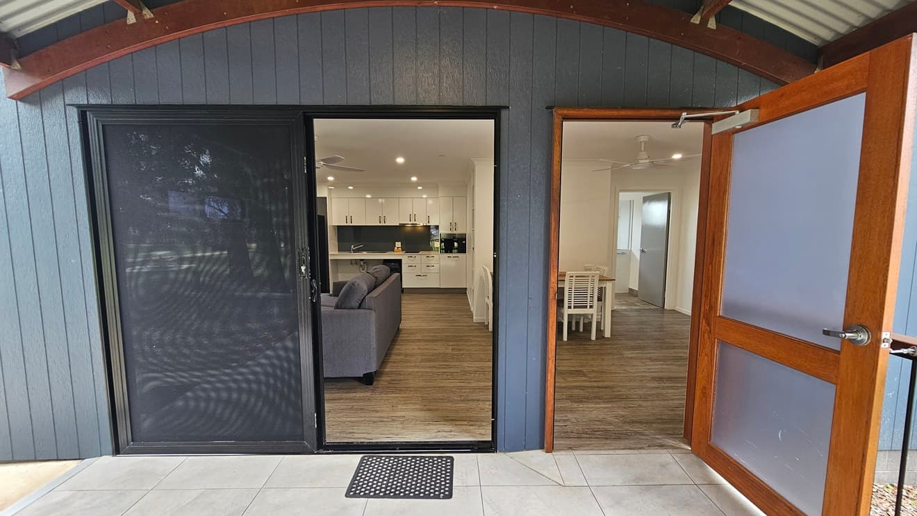 There are two entrance doors. The right one is a manual sliding glass door, equipped with a non-slippery mat while the left door is a manual outward opening door. Inside, the floor is timber, and the walls and ceiling are white with ceiling fans installed. The kitchen area, dining table, and a grey couch are also visible. Outside, the entrance features an arched ceiling with a corrugated roof, and the wall has grey wooden planks. The floor has grey tiles.