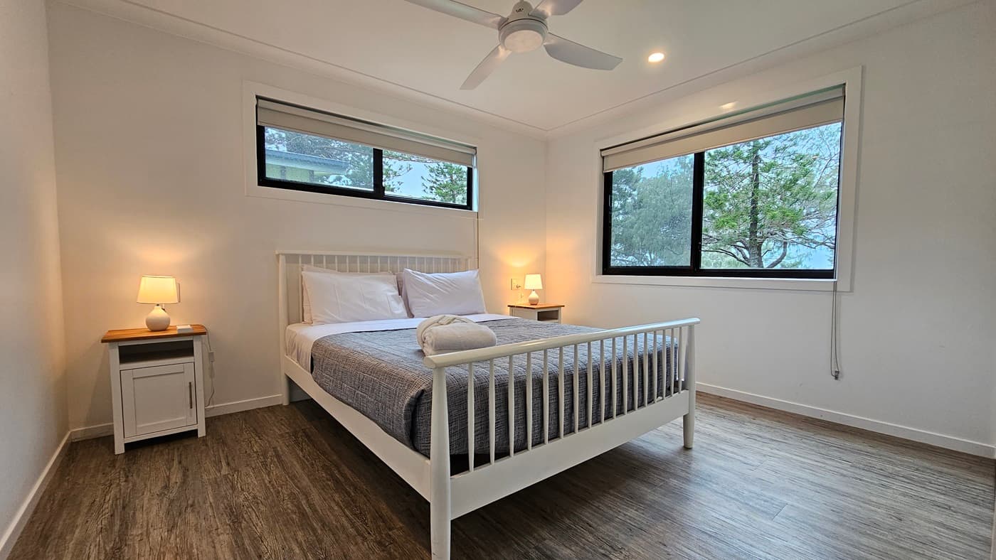 In the centre stands a white queen-size bed adorned with a grey cover. White pillows and a neatly rolled white towel rest atop. Flanking the bed are two bedside tables, each with a lampshade. A window is situated on the central and right walls. The walls and ceiling are painted white, illuminated by LED lights and a ceiling fan. The floor is timber.