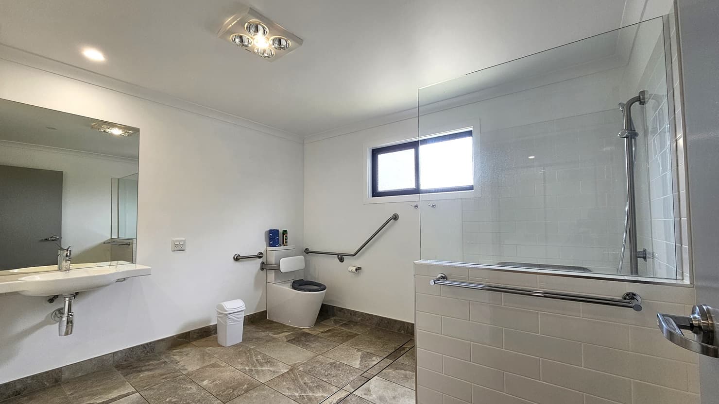 In the accessible bathroom, a toilet bowl with hand grabs is situated in the left corner, accompanied by a rubbish bin and a tissue dispenser. Adjacent to this is a hand basin with a mirror mounted on the glass wall. To the right, a shower is present, separated by a combination of glass and grey brick tiles. The floor features grey tiles with a marble design. The walls are white, and the ceiling is white with LED lights.