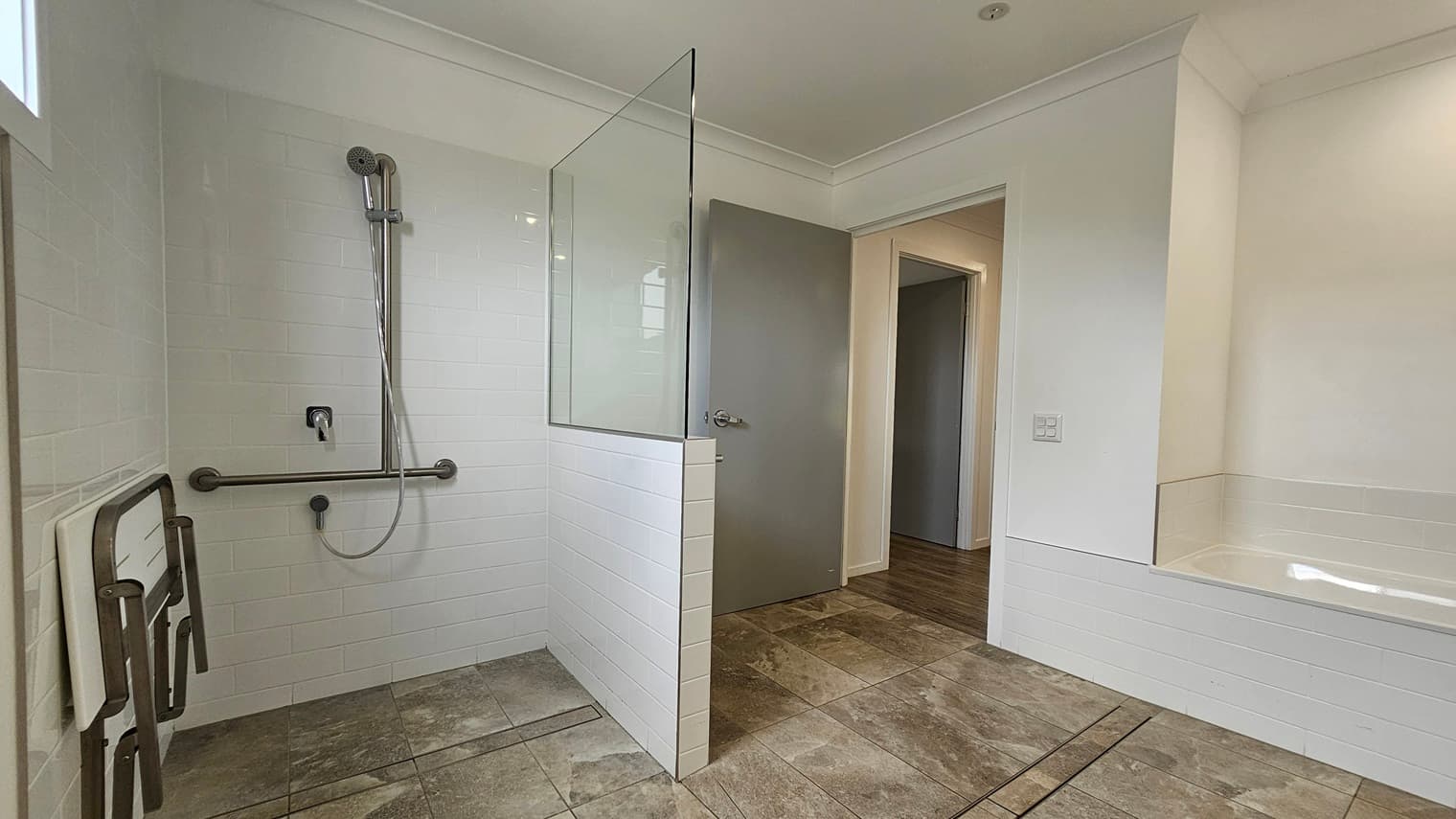 In the accessible bathroom, a shower with a handrail and foldable bench is on the left, separated by a partition of glass and grey brick tiles. On the right is a bath tub against the wall. The floor boasts grey tiles with a marble design. The walls and the ceiling are white with LED lights.