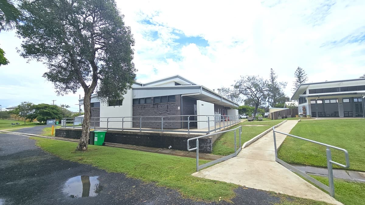 In a grassy area stands a low-rise facility with a grey brick facade. A brick pathway leads to it, alongside a step-free ramp with railings. Four gas tanks are displayed in front of the building. To the left is a round circular structure, and to the right is a wooden planter box with plants. Trees surround the area.