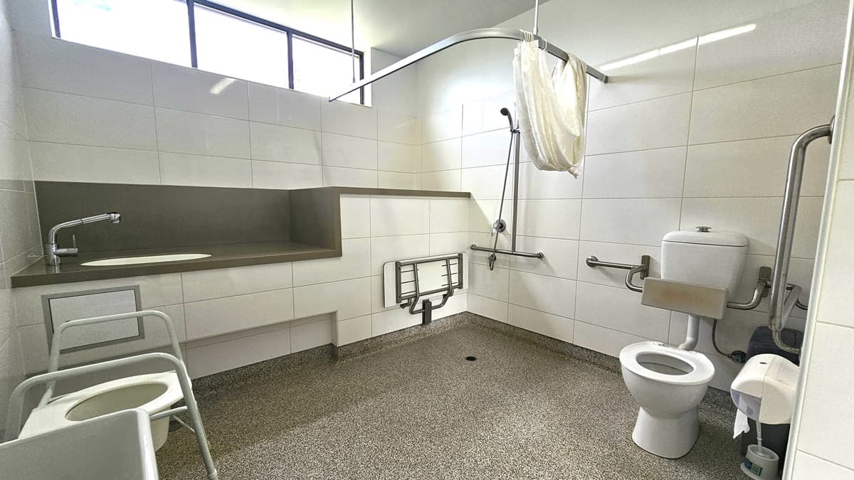 Inside the accessible bathroom, a toilet bowl with hand grabs, a tissue dispenser, a rubbish bin, and a toilet brush are on the right. On the left is a shower with handrails and curtain and a foldable bench chair mounted to the wall. Adjacent, is a lavatory with a sink. The walls feature white tiles with glass windows. A toilet pot is positioned on the left, near a rubbish bin. Floor tiles have a granite design.