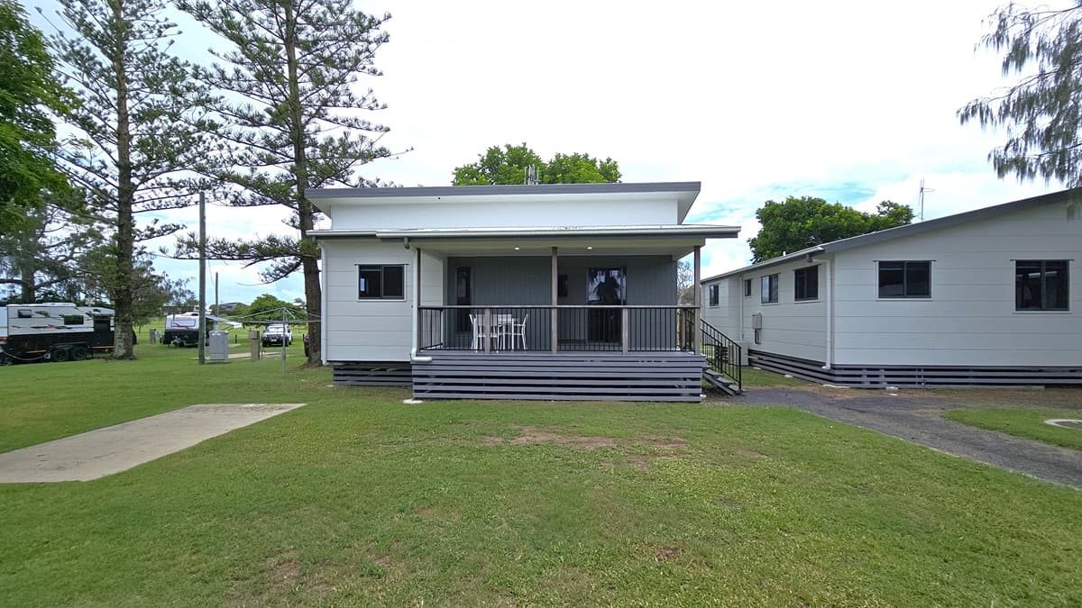 The standard cabin is a one-storey structure with a verandah, featuring steps with handrails on the right. Its exterior displays white and grey walls with pitched roofing. Tables and chairs are placed on the veranda, while another cabin stands to the right. Surrounding the area are green grass and pine trees, with trucks and vehicles visible under the shade of trees.