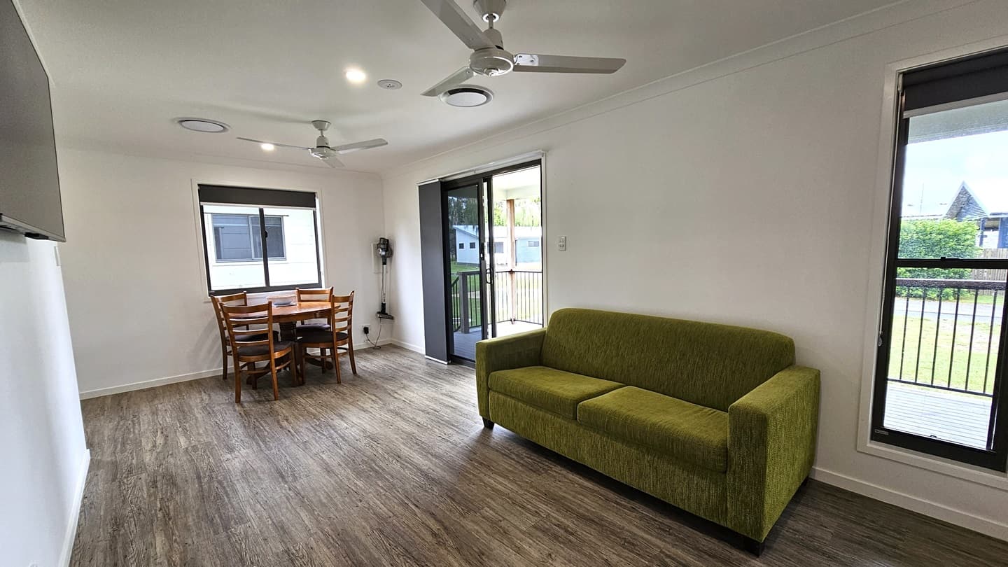 To the right is a glass sliding door leading to the verandah, alongside another door. A green couch rests against the middle wall, with a flatscreen TV mounted on it. A round wooden dining table sits near a glass window. The floor is timber, the walls are white, and the ceiling features ceiling fans and LED lights. Outside, green grass and trimmed shrubs are visible.