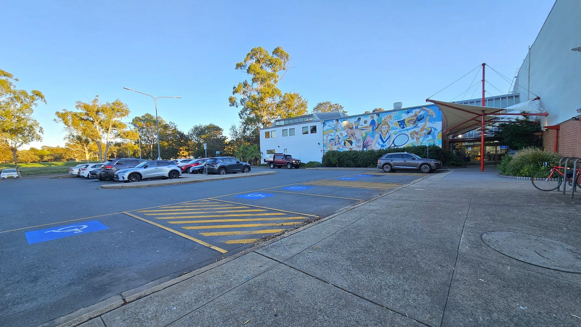 A car park with several vehicles on the left, including a silver car in the foreground. The ground has marked disabled parking spaces with blue and yellow lines. In the centre, a building with a colourful mural depicting people engaged in various activities. On the right, a covered entrance with red structural elements and a bicycle parked nearby. Trees and greenery are visible in the background. The pavement is concrete.