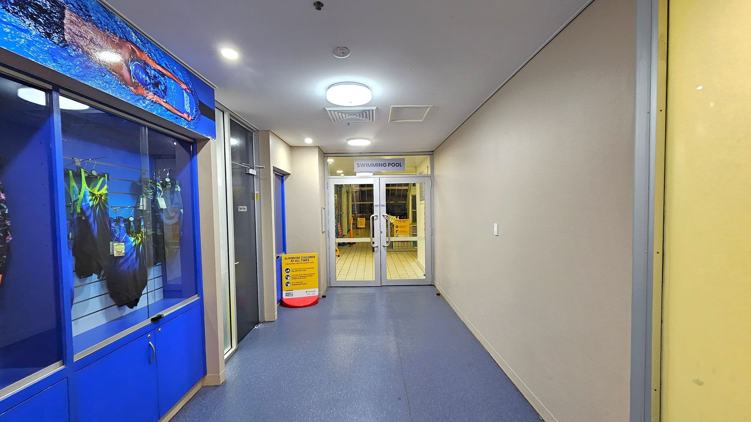 A corridor with a blue floor and a white ceiling with circular lights. On the left, there is a display case with swimming costumes and a large image of a swimmer above it. On the right, a plain wall. At the end, glass doors with a sign above reading "SWIMMING POOL". A yellow caution sign is visible near the doors.