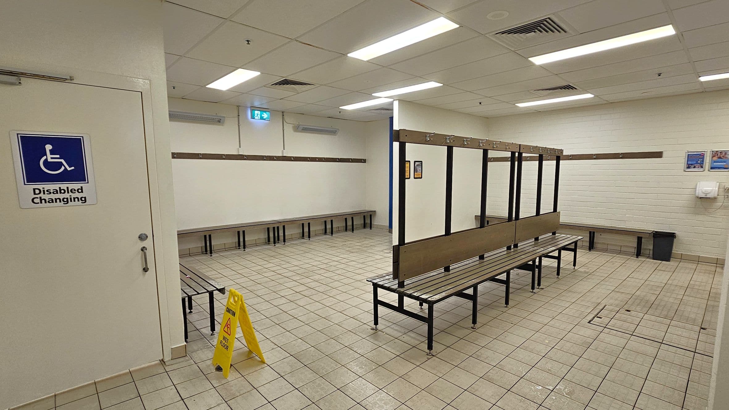A changing room with tiled flooring and a suspended ceiling with fluorescent lights. On the left, a door marked "Disabled Changing" is visible. In the centre, there are benches with hooks above them for hanging clothes. The right side has more benches and a waste bin. A yellow caution sign indicating a wet floor is placed near the centre. The walls are light-coloured with a few posters and a hand dryer mounted on the right wall.