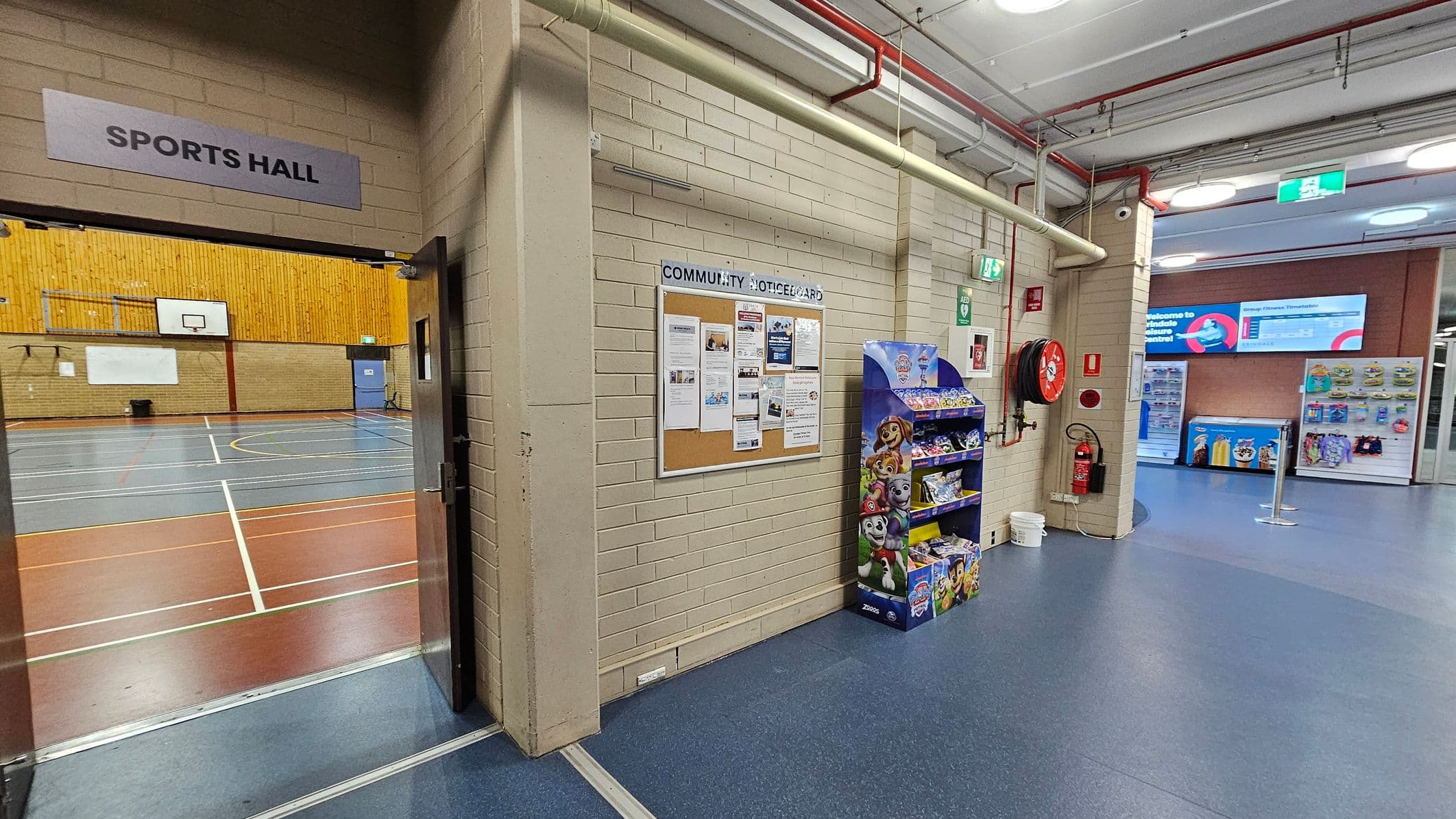 On the left, an open door leads to a sports hall with a wooden wall and basketball hoops. In the centre, a community noticeboard is mounted on a brick wall, with various papers pinned to it. Below, a display stand with children's snacks is visible. On the right, a fire hose reel and extinguisher are mounted on the wall. The floor is blue, and the ceiling has exposed pipes and lights. In the background, vending machines and a digital sign are visible.