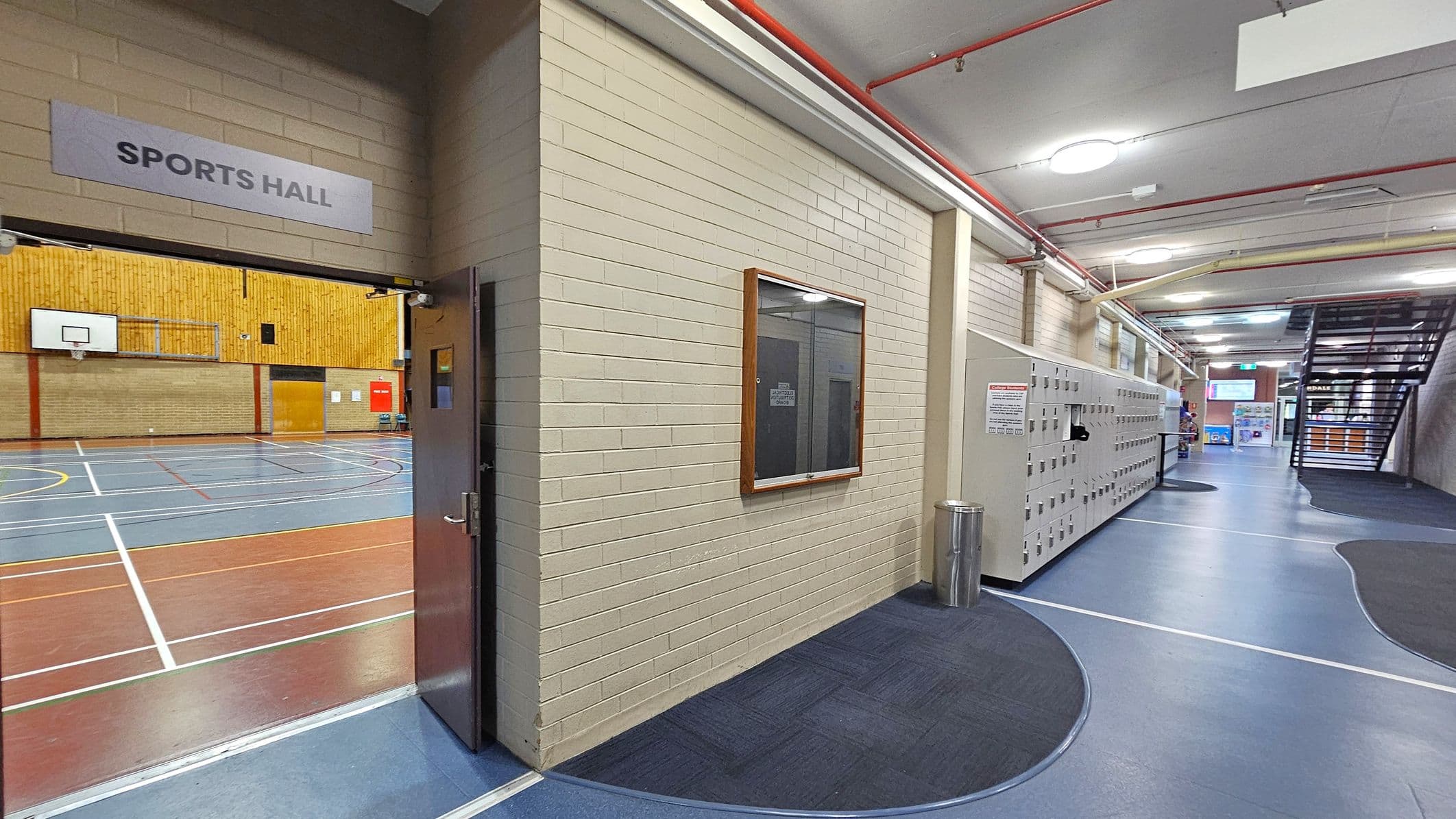 A corridor with a sports hall entrance on the left, marked by a sign above the door. Inside, a basketball hoop and court lines are visible. The right side features a row of lockers along a blue and grey floor. A notice board is mounted on the wall near a metal bin. The ceiling has exposed pipes and circular lights. In the background, a staircase leads upwards, and there are posters and signs near the exit.