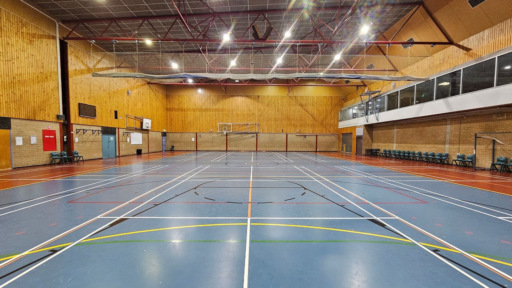 A spacious indoor sports hall with a blue floor marked with various sports lines in white, yellow, and orange. The walls are clad in light wood panelling. On the left, there are chairs and a red fire hose reel. The right side features a row of chairs against a brick wall and a large windowed viewing area above. The ceiling is high with exposed beams and lighting fixtures. A basketball hoop is visible at the far end.