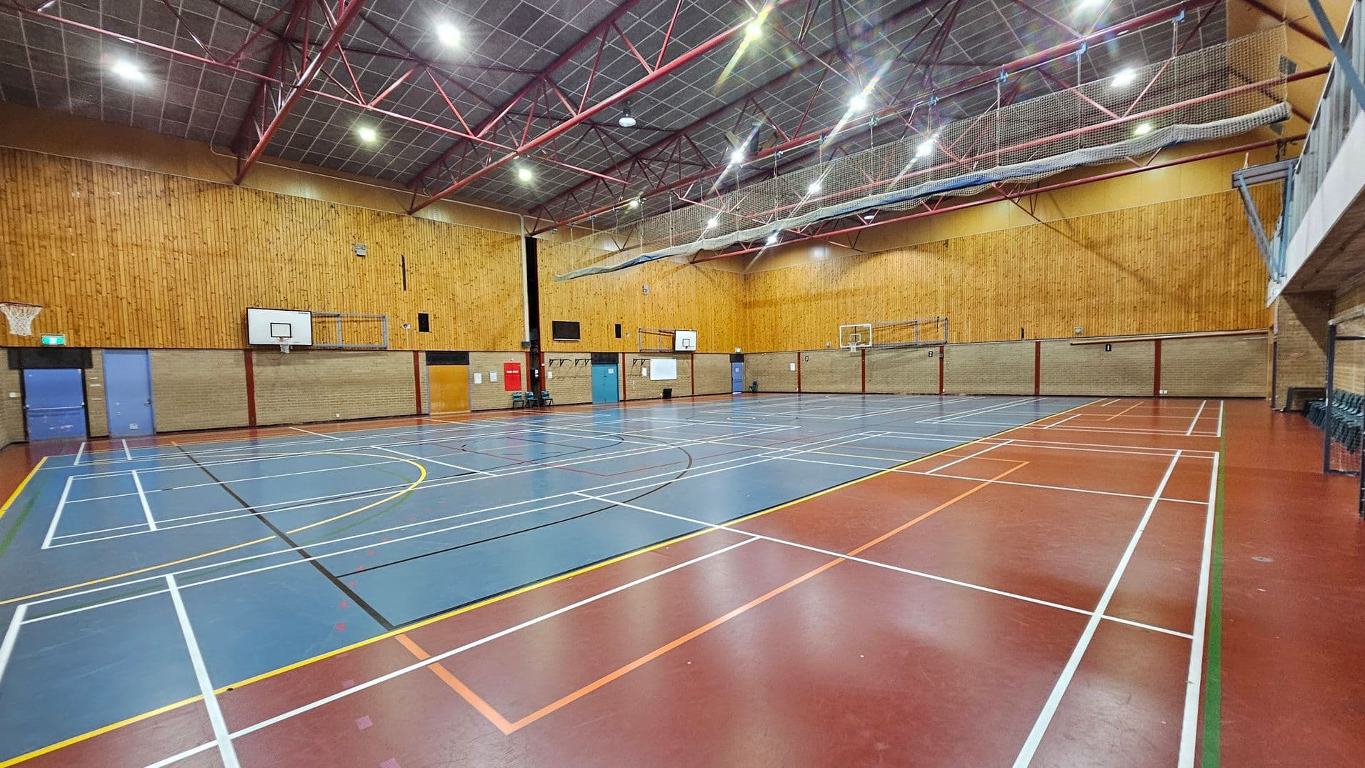 A spacious indoor sports hall with a high ceiling supported by red metal beams. The floor is marked with various sports lines in white, yellow, and orange on a blue and red surface. On the left, there are basketball hoops and blue doors. The walls are a mix of brick and wood panelling. On the right, a row of stacked chairs is visible. The ceiling features bright overhead lights and a netting system.