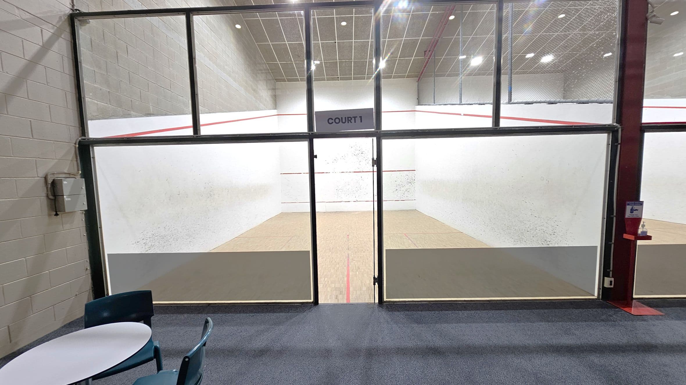 A squash court is visible through large glass doors labelled "Court 1". The court has white walls with red boundary lines and a wooden floor. In the foreground, there is a small round table with blue chairs on a carpeted floor. The ceiling is high with a grid pattern and bright lights. The left wall is made of grey bricks, and there is a hand sanitiser dispenser mounted on the right side near the entrance.