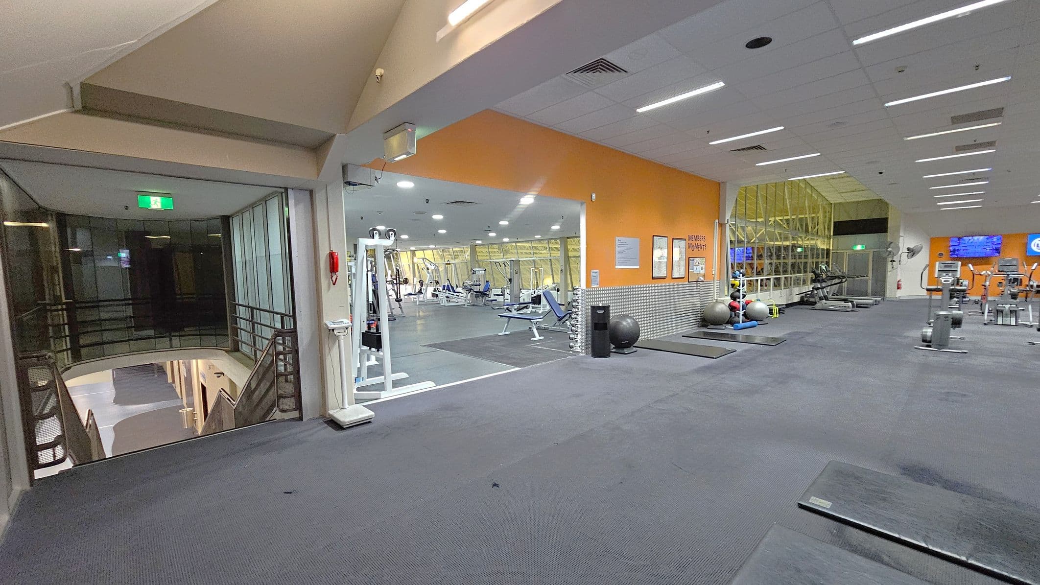 A gym interior with a grey carpeted floor and a white ceiling with recessed lighting. On the left, there's a glass-walled area with a staircase leading down. In the centre, an open space with various gym equipment, including weight machines and benches, is visible. The right side features an orange wall with posters, exercise balls, and more gym equipment like treadmills. A large window with a view of the outside is in the background.