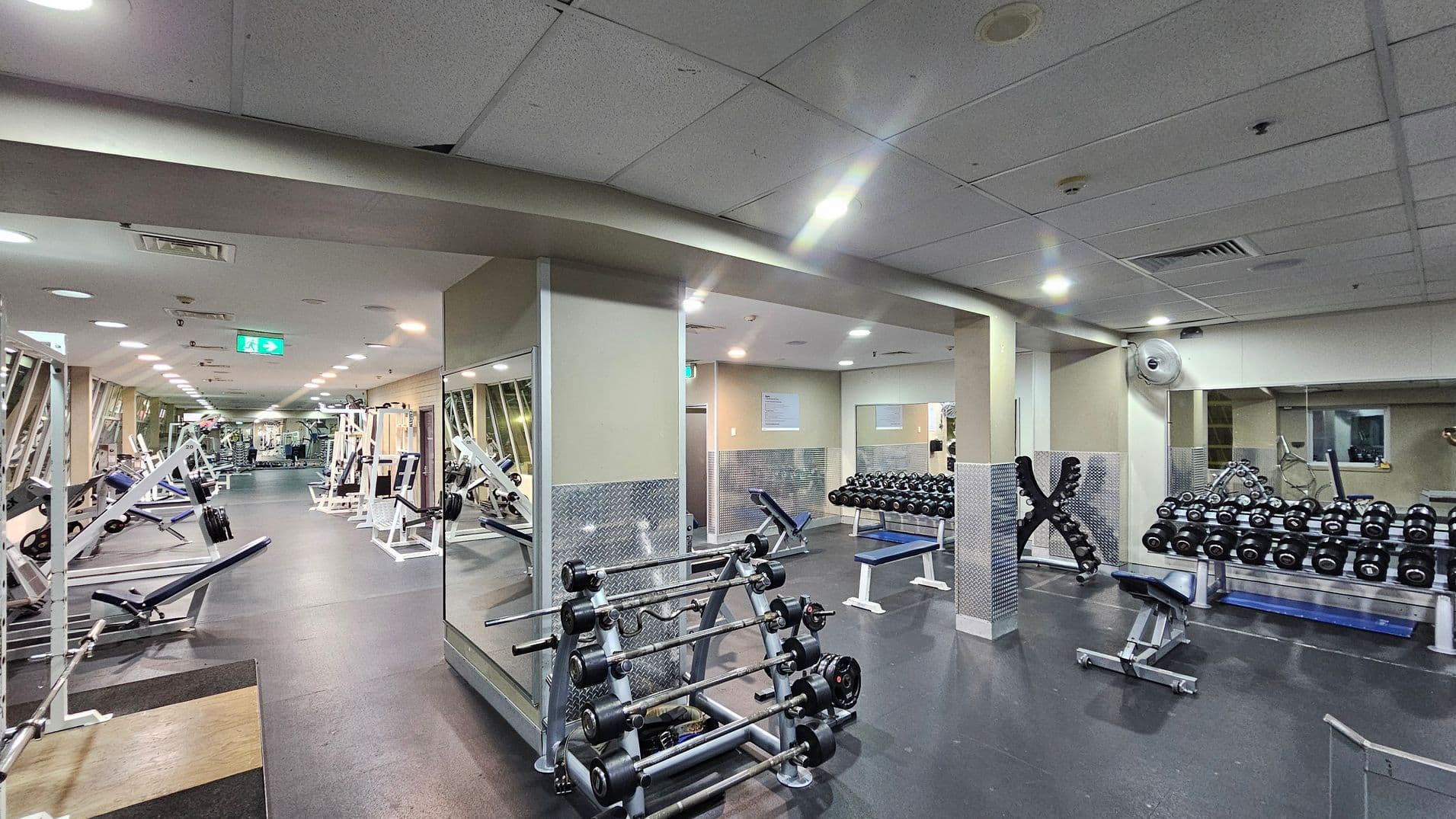 A spacious gym with a variety of equipment. On the left, there are weightlifting benches and machines with weights. In the centre, a rack holds multiple barbells. On the right, there are rows of dumbbells on racks and a bench. The floor is dark and rubberised, while the ceiling is white with recessed lighting. Mirrors line the walls, enhancing the sense of space. An emergency exit sign is visible in the background.