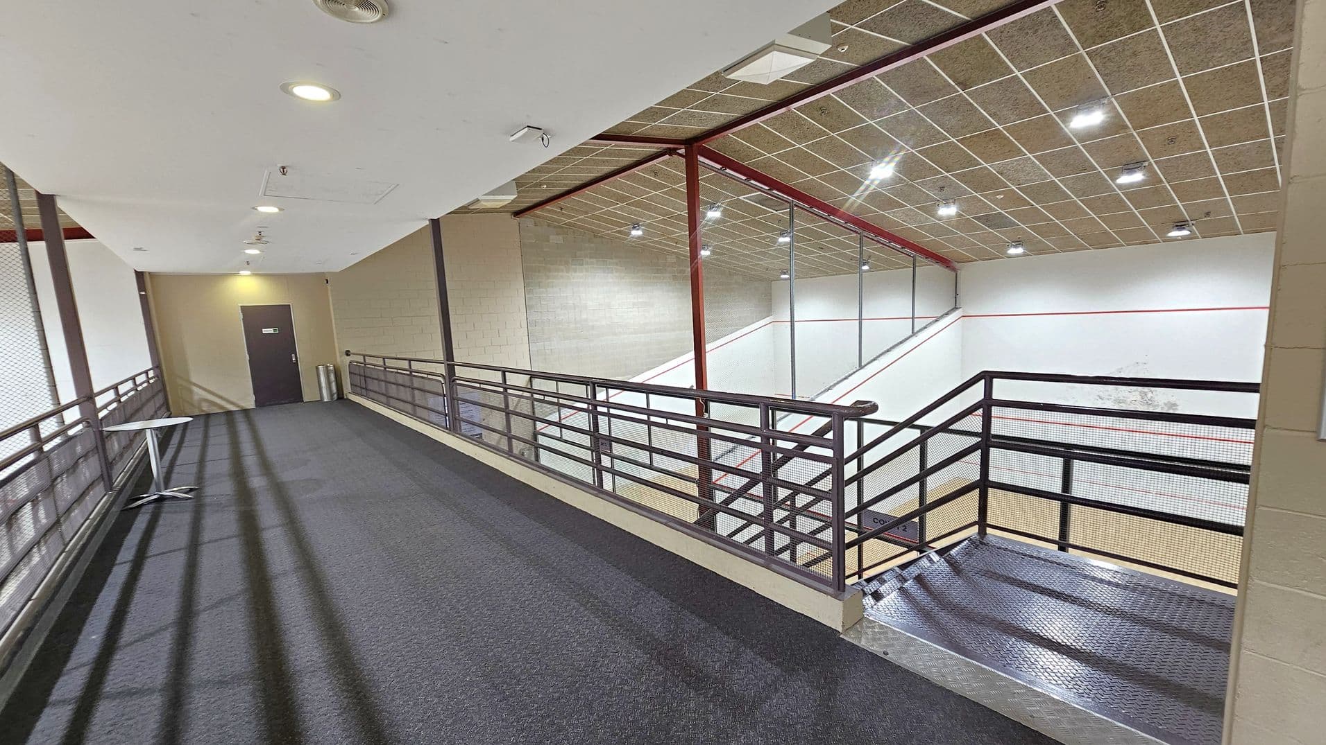 A corridor with a dark carpeted floor and a white ceiling with recessed lights. On the left, there's a small table and a door at the end of the corridor. The right side features a metal railing overlooking a squash court below, with a wooden floor and white walls marked with red lines. The ceiling above the court is tiled with multiple lights.