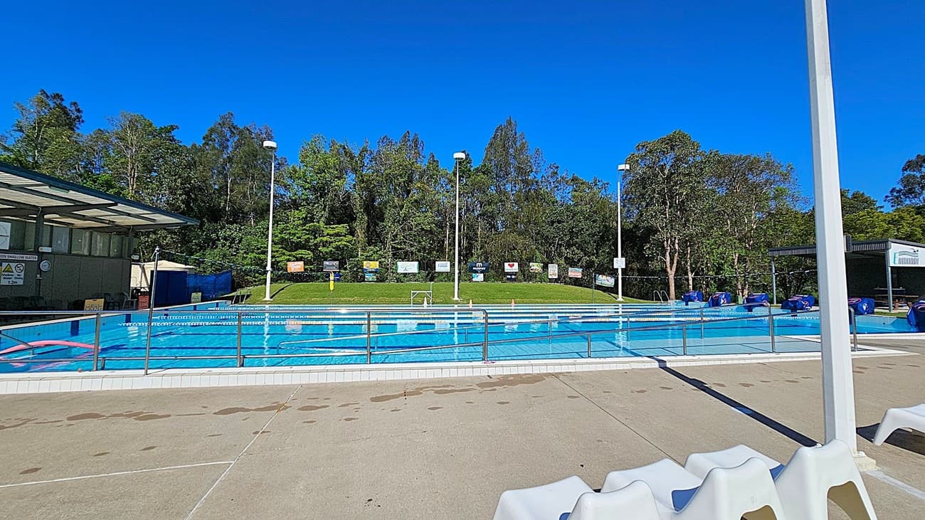 Outdoor swimming pool with clear blue water, surrounded by a metal railing. On the left, there is a covered area with signs and a small building. In the middle, the pool is divided into lanes with floating lane markers. The background features a grassy hill with various signs and tall trees. On the right, there are several blue pool covers and another small structure. The floor around the pool is concrete with some water spots.