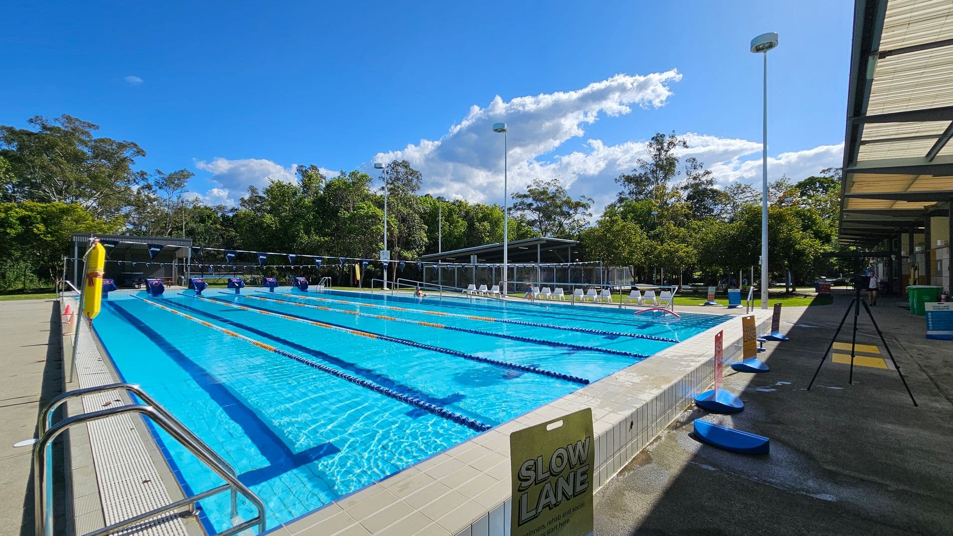 An outdoor swimming pool with multiple lanes, each marked by blue and yellow lane dividers. On the left, there is a metal handrail and a sign indicating the "Slow Lane." The pool deck is tiled. On the right, there is a covered walkway with a tripod and various signs. In the background, there are trees and a shaded seating area with white chairs. Tall light poles are positioned around the pool area. The floor is concrete, and the ceiling of the walkway is made of translucent panels.
