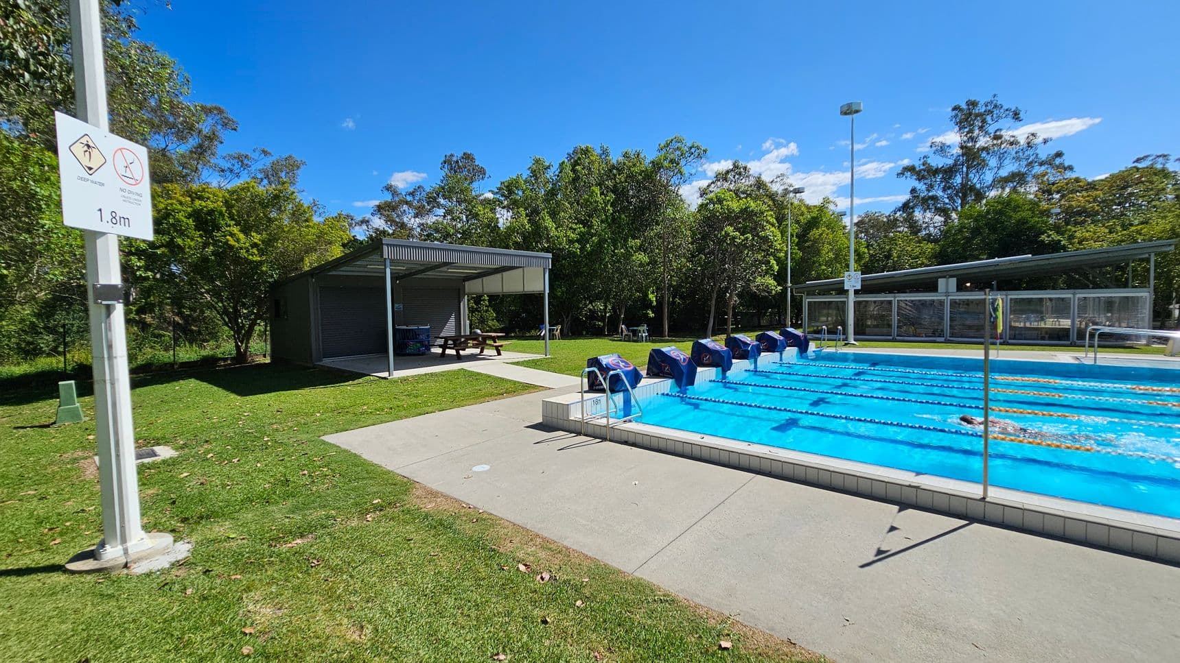 Outdoor swimming pool with lanes on the right, covered by blue lane markers. A swimmer is in one of the lanes. On the left, a sign on a pole indicates "Deep Water" and "No Diving" with a depth of 1.8m. Behind the sign, there is a shaded seating area with picnic tables. The background features green grass, trees, and a fence. The foreground has a concrete pool deck and grassy area.