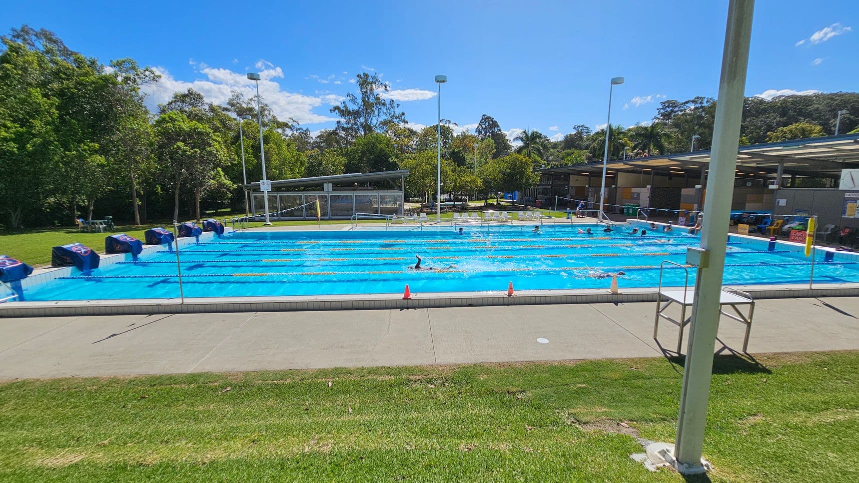 An outdoor swimming pool with multiple lanes, each marked by lane ropes. Swimmers are in the pool, and starting blocks are positioned at one end. On the left, there is a grassy area with trees and some chairs. On the right, there is a covered area with seating and various facilities. The pool deck is concrete, and there are a few orange cones placed along the edge. A tall light pole is in the foreground on the right side.