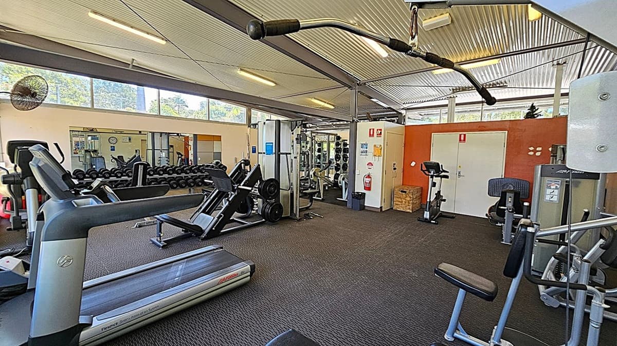 A gym with various exercise equipment. On the left, there are treadmills and a fan mounted on the wall. In the middle, there are rows of dumbbells and a leg press machine. On the right, there are stationary bikes, a fire extinguisher, and a door with an orange wall behind it. The ceiling is corrugated metal with fluorescent lights, and the floor is carpeted. Large windows near the ceiling let in natural light.