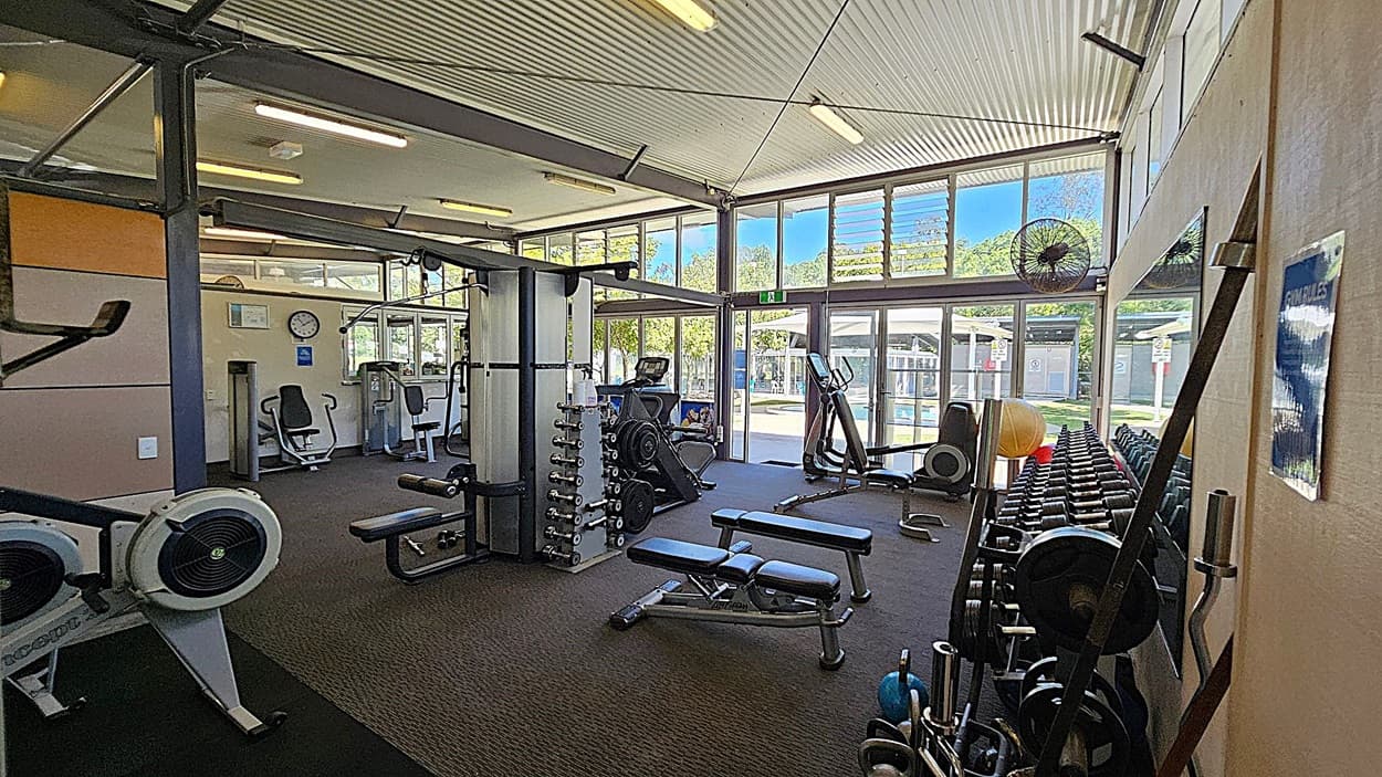 A gym with various exercise equipment. On the left, there are rowing machines and a seated exercise machine. In the middle, there is a multi-functional weight machine, benches, and a rack of dumbbells. On the right, there are more weights, exercise balls, and a large mirror. The background features large windows and glass doors, allowing natural light to enter. The ceiling is corrugated with fluorescent lights, and the floor is carpeted.