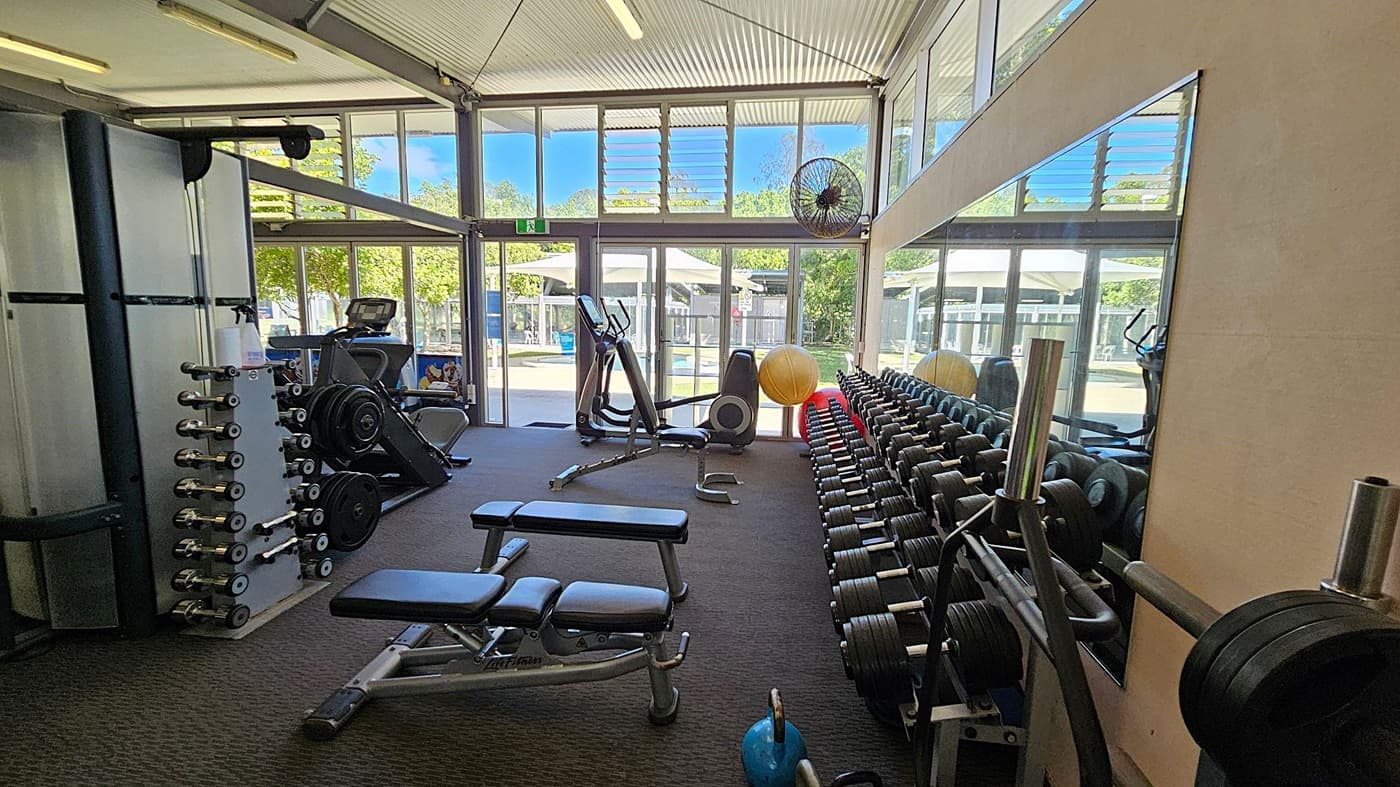 A gym with large windows and glass doors allowing natural light to enter. On the left, there is a weight machine and a rack of dumbbells. In the middle, there are two adjustable benches and a stationary bike. On the right, there is a long mirror reflecting the equipment and a row of dumbbells. A large yellow exercise ball and a blue kettlebell are also visible. The floor is carpeted, and the ceiling has exposed beams with fluorescent lights.