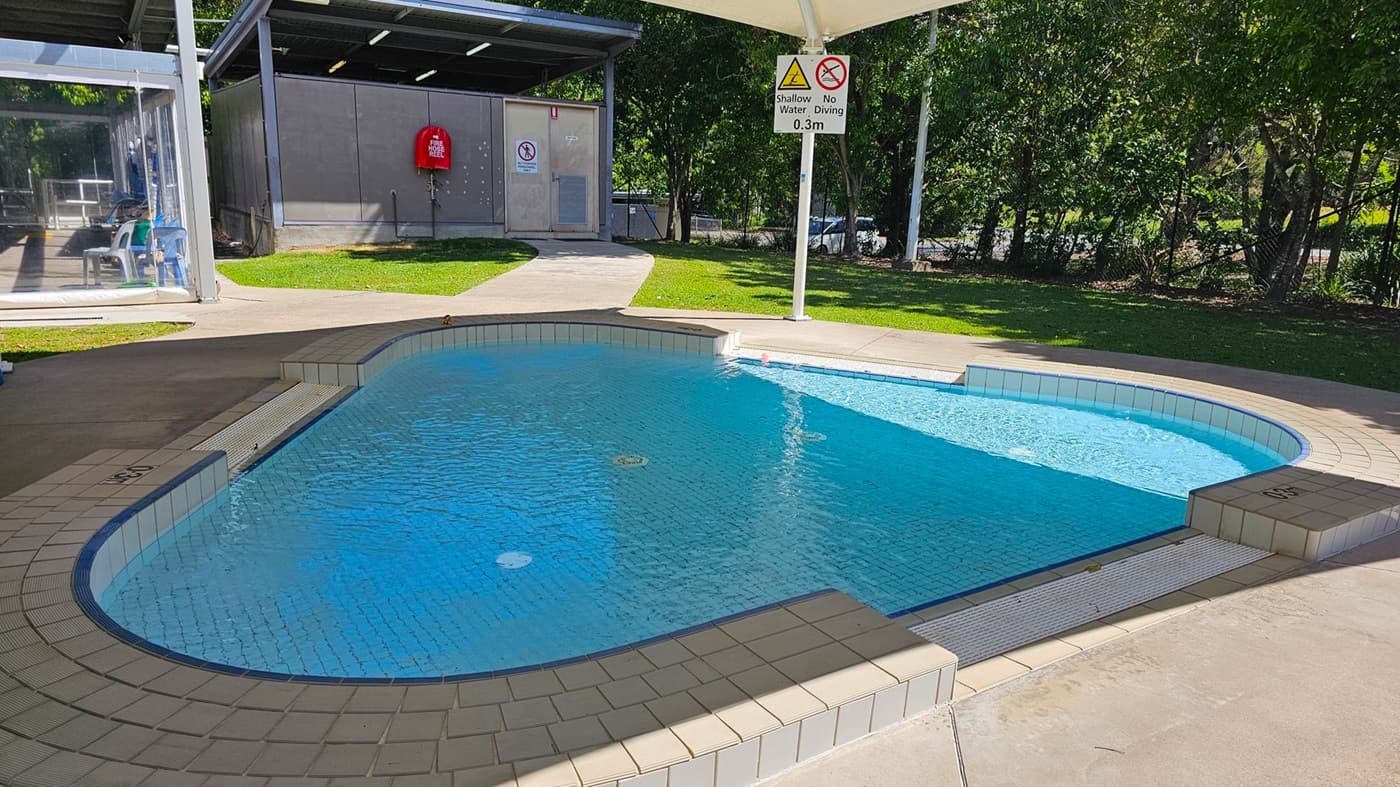 A small, circular wading pool is in the centre, covered by a white canopy supported by four poles. Surrounding the pool is a concrete pathway, with grass and trees on either side. To the left, there is a glass-walled building with chairs inside and a red fire extinguisher mounted on the exterior wall. In the foreground, a blue sign with "Aquatic Supervision" guidelines is visible. To the right, there are a few plastic chairs and a fence with parked cars behind it.