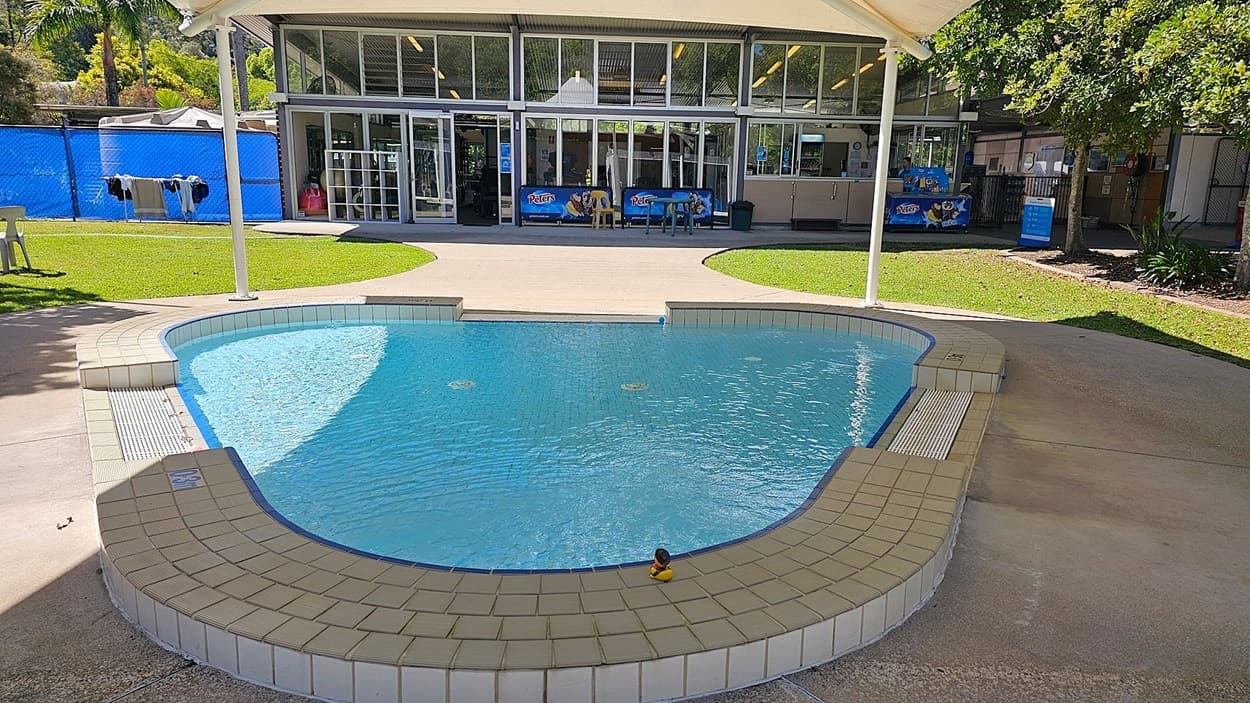 A small, shallow swimming pool with a tiled edge and a rubber duck floating in the water. The pool is surrounded by a concrete walkway. In the background, there is a building with large glass windows and doors, displaying signs for "Peters" ice cream. To the left, there is a grassy area with a white plastic chair and clothes hanging on a line. To the right, there are trees and more grassy areas.