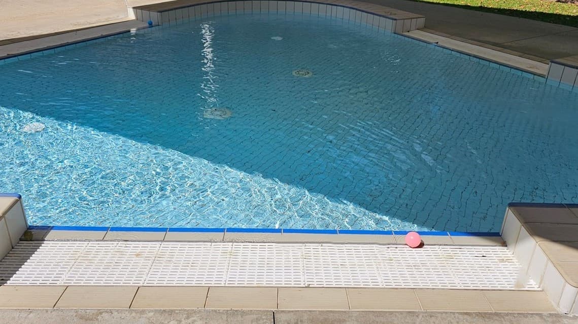 A swimming pool with clear blue water. The pool has a tiled edge and a shallow end marked "0.3m". On the poolside, there are a few leaves and a pink ball. The surrounding area includes a concrete walkway and a grassy area in the background. The pool is partially shaded by a structure.