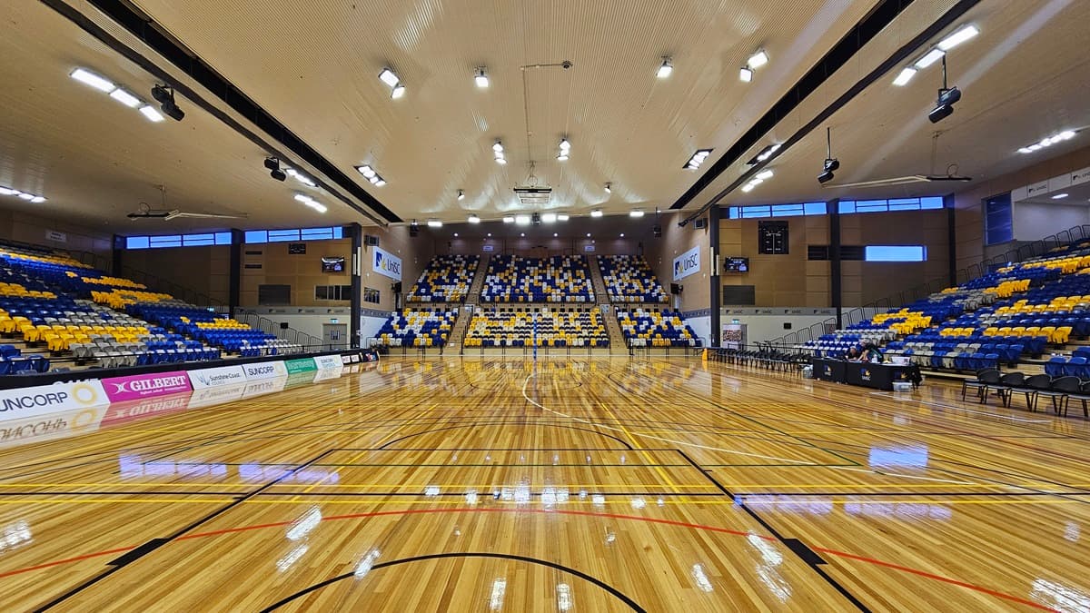 On the left, there are rows of blue and yellow seats with advertising banners along the lower section. The middle features a large seating area with blue, yellow, and white seats arranged in a pattern, and a basketball court with polished wooden flooring. The right side has more seating in blue and yellow, with additional advertising banners. The foreground shows the basketball court lines and the ceiling is high with recessed lighting and ventilation fans.