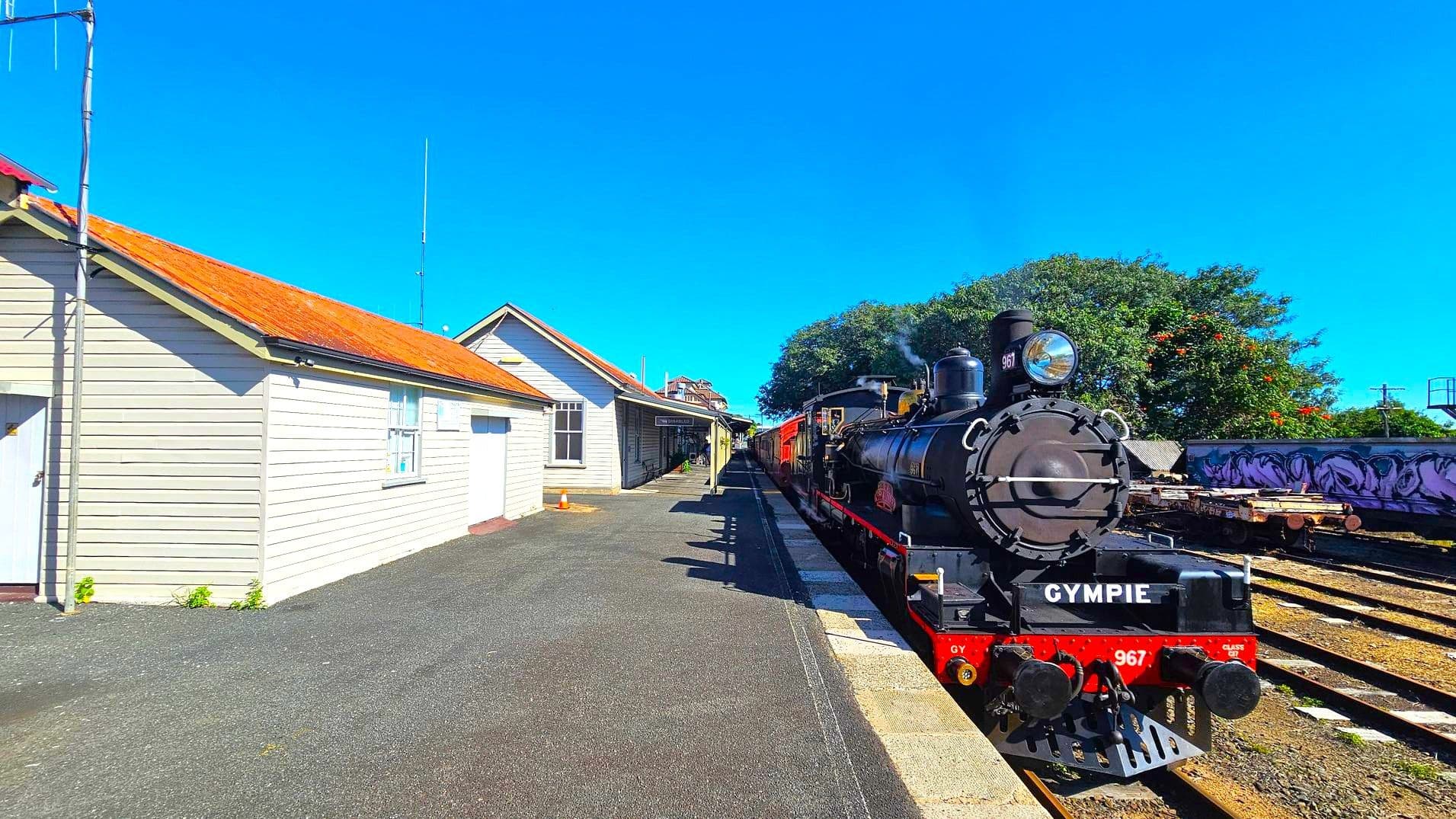 A vintage steam locomotive is on the right, labelled "GYMPIE" with the number 967. It is stationed on railway tracks beside a platform. On the left, there is a light-coloured wooden building with a red roof, featuring windows and a door. The platform is paved, and a small orange traffic cone is visible near the building. In the background, there are trees and additional railway tracks with some graffiti on a wall.