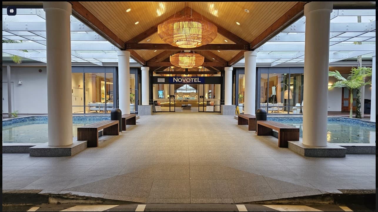 On the left, there is a small pool bordered by a low stone wall, with greenery visible around it. A wooden bench sits beside the pool. To the right, another wooden bench is positioned near the entrance, which features large glass doors displaying the "Novotel" sign. The floor is tiled in light grey, and the ceiling is wooden with exposed beams, complemented by large, decorative light fixtures.
